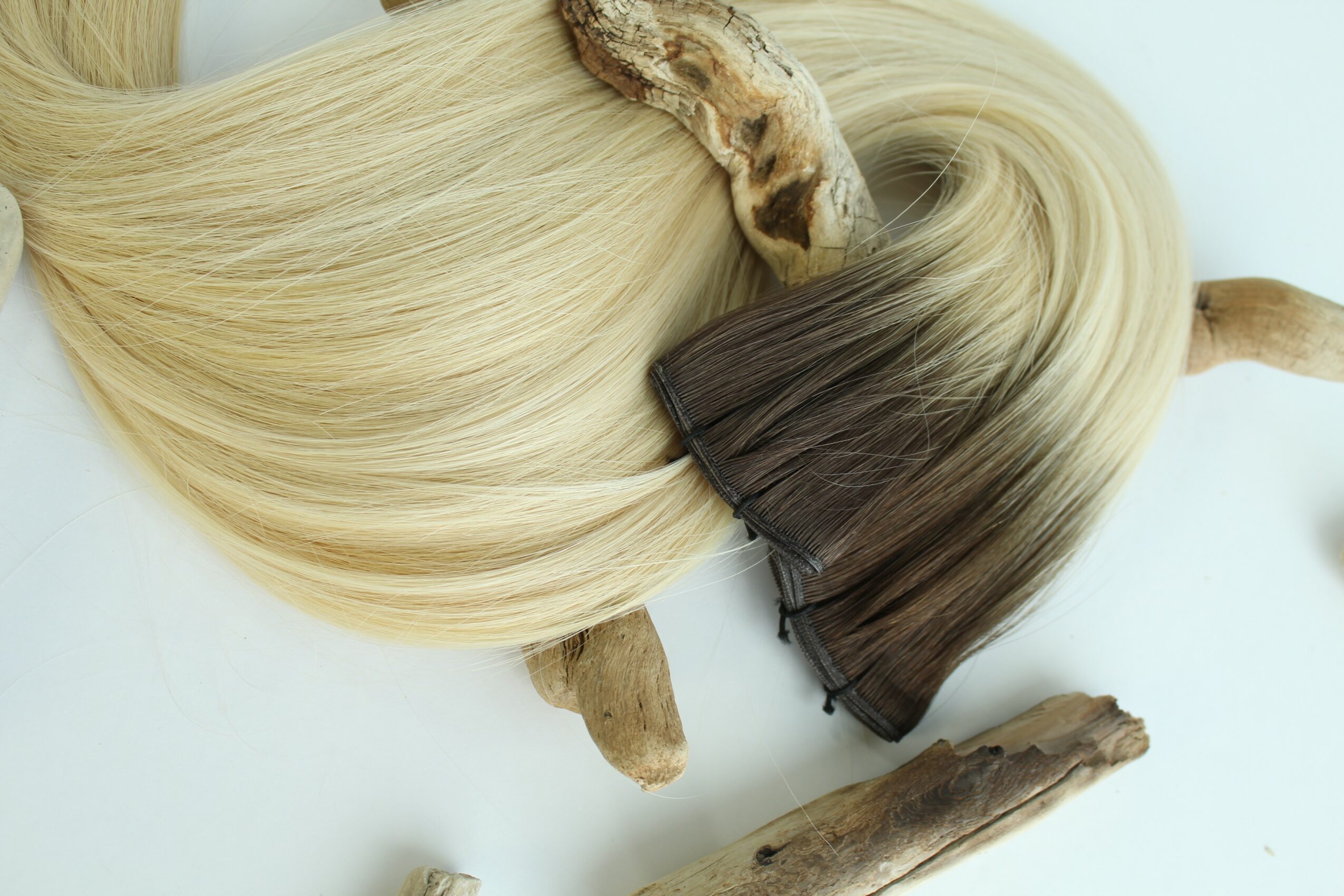 Blonde and brown hair extensions with natural roots, placed on a white surface, with a piece of wood nearby.