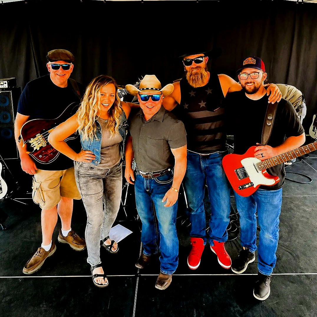 Group of five people posing with musical instruments on stage, smiling, black background, casual clothing, sunglasses, and hats.