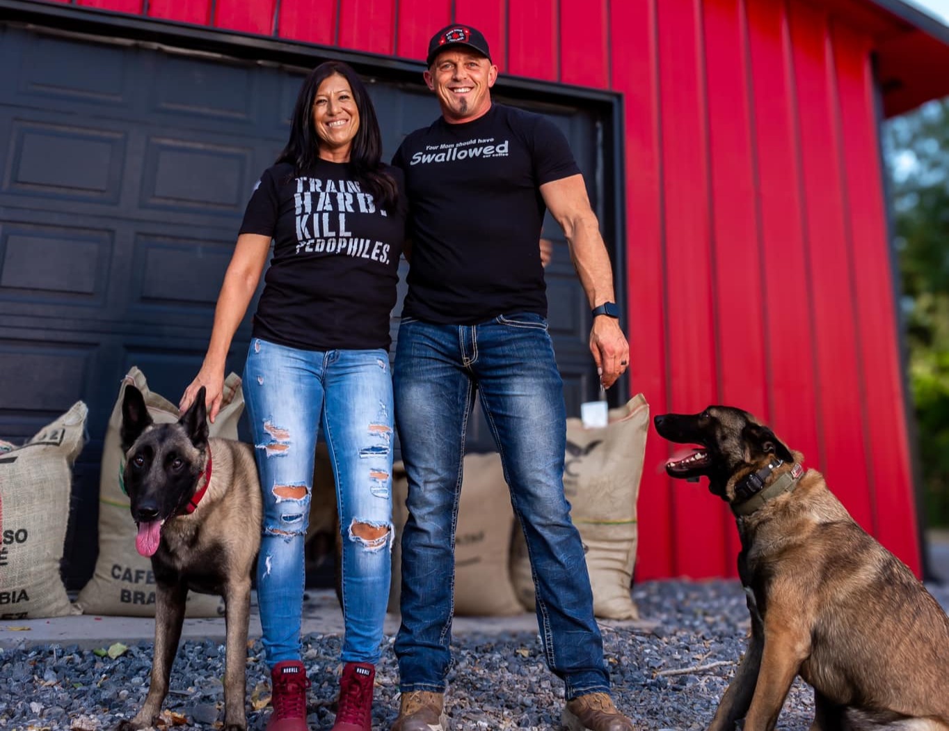 Two people stand outside a red building with two dogs, one German Shepherd and one Belgian Malinois, on a gravel surface.