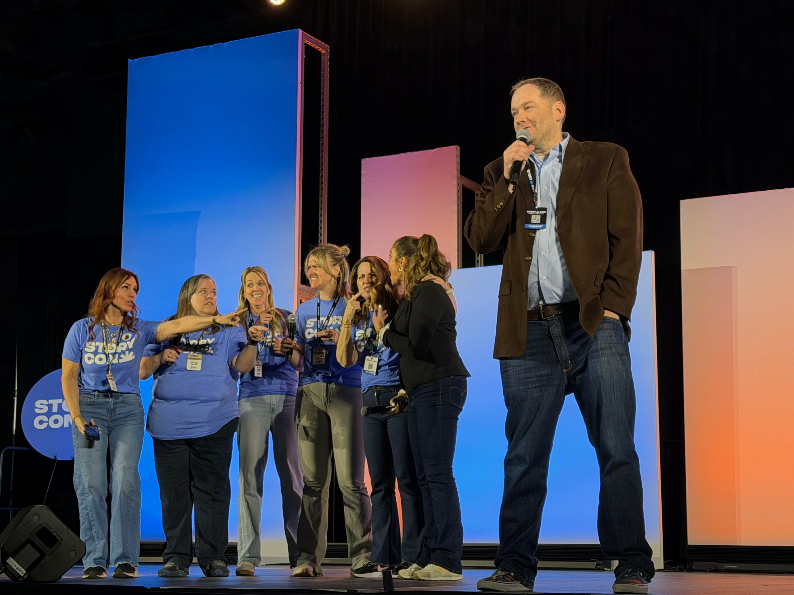 Man speaking into microphone on stage with group of women in blue shirts, colorful background panels, and dark curtain.