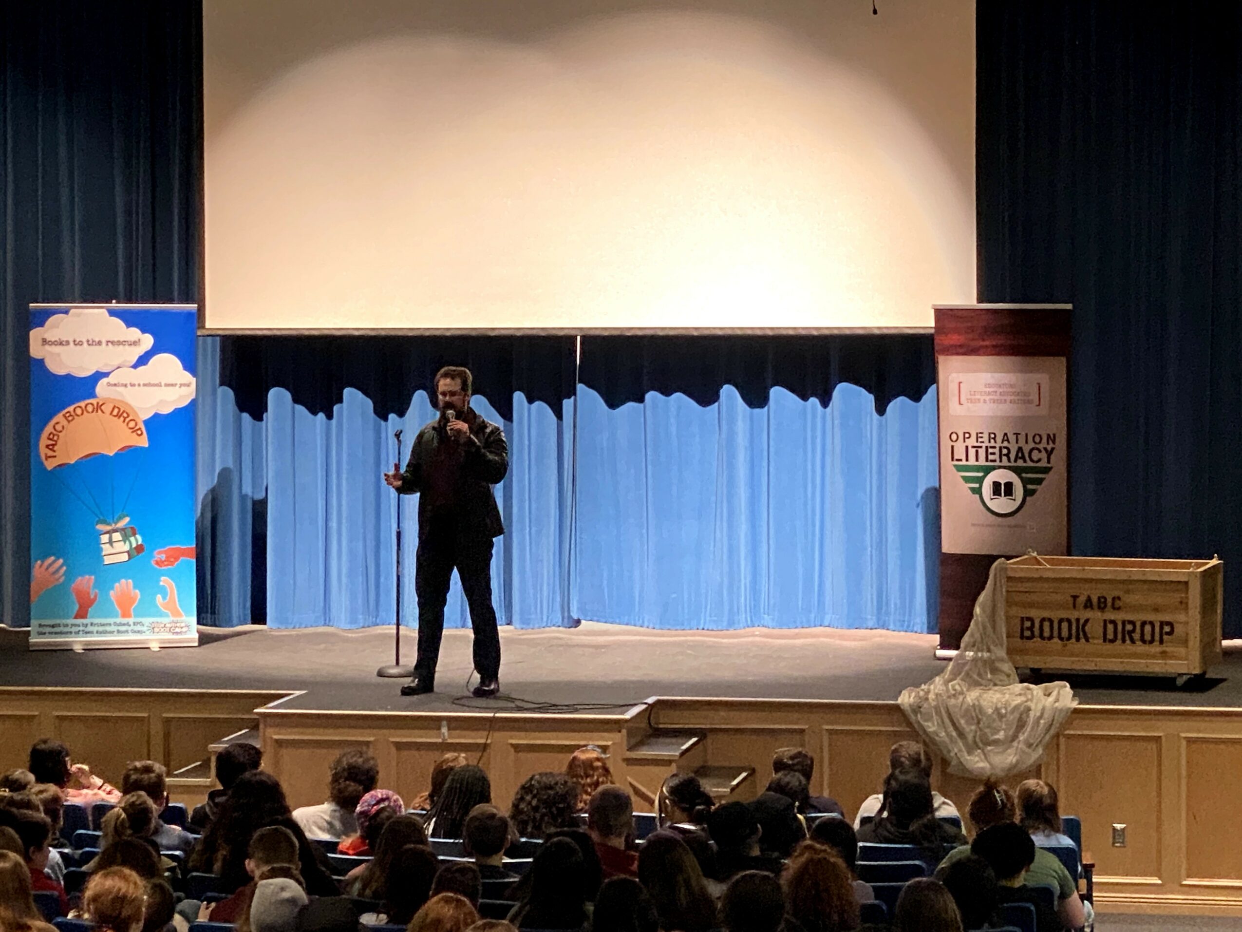 Person standing on stage speaking to an audience with banners and a box on stage, audience seated in front.