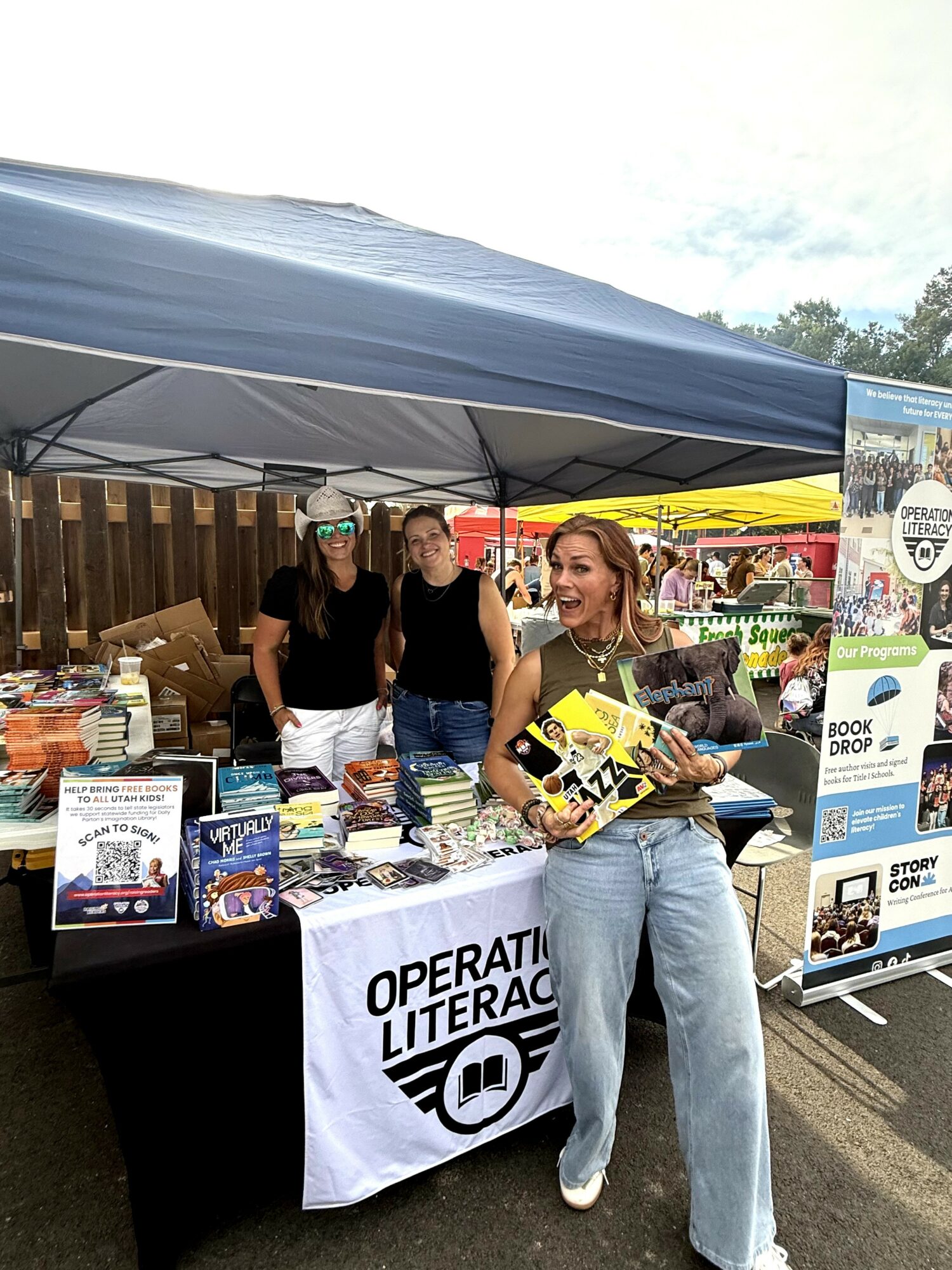 Woman holding books and smiling at outdoor literacy event booth with three people behind table, under canopy, with books displayed.
