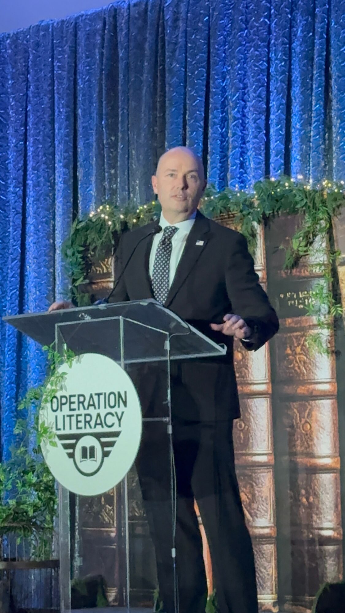Man in suit speaking at a podium with Operation Literacy logo, blue curtain backdrop, and greenery.