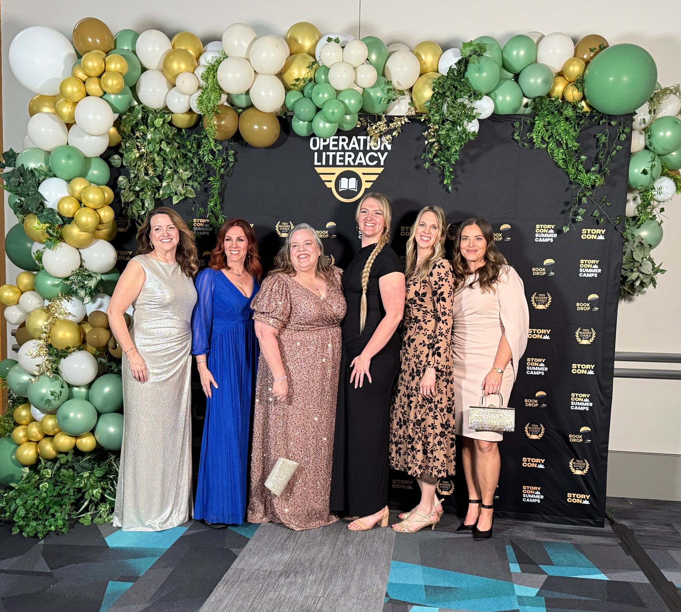 Six women in formal dresses stand in front of a decorated backdrop with balloons and greenery at an event.