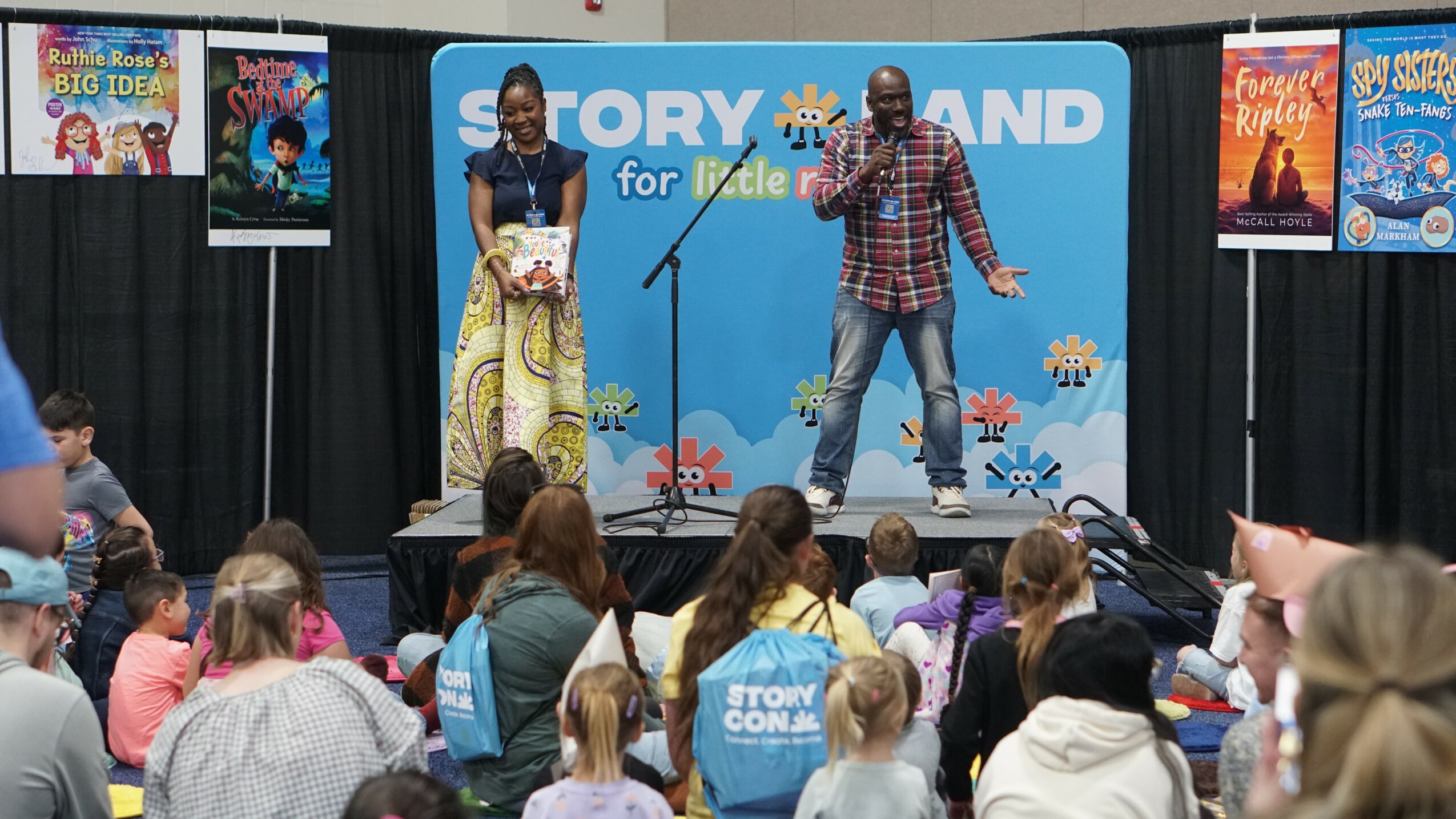 Two presenters on stage with a blue backdrop, speaking to an audience of children and adults, colorful posters on sides.