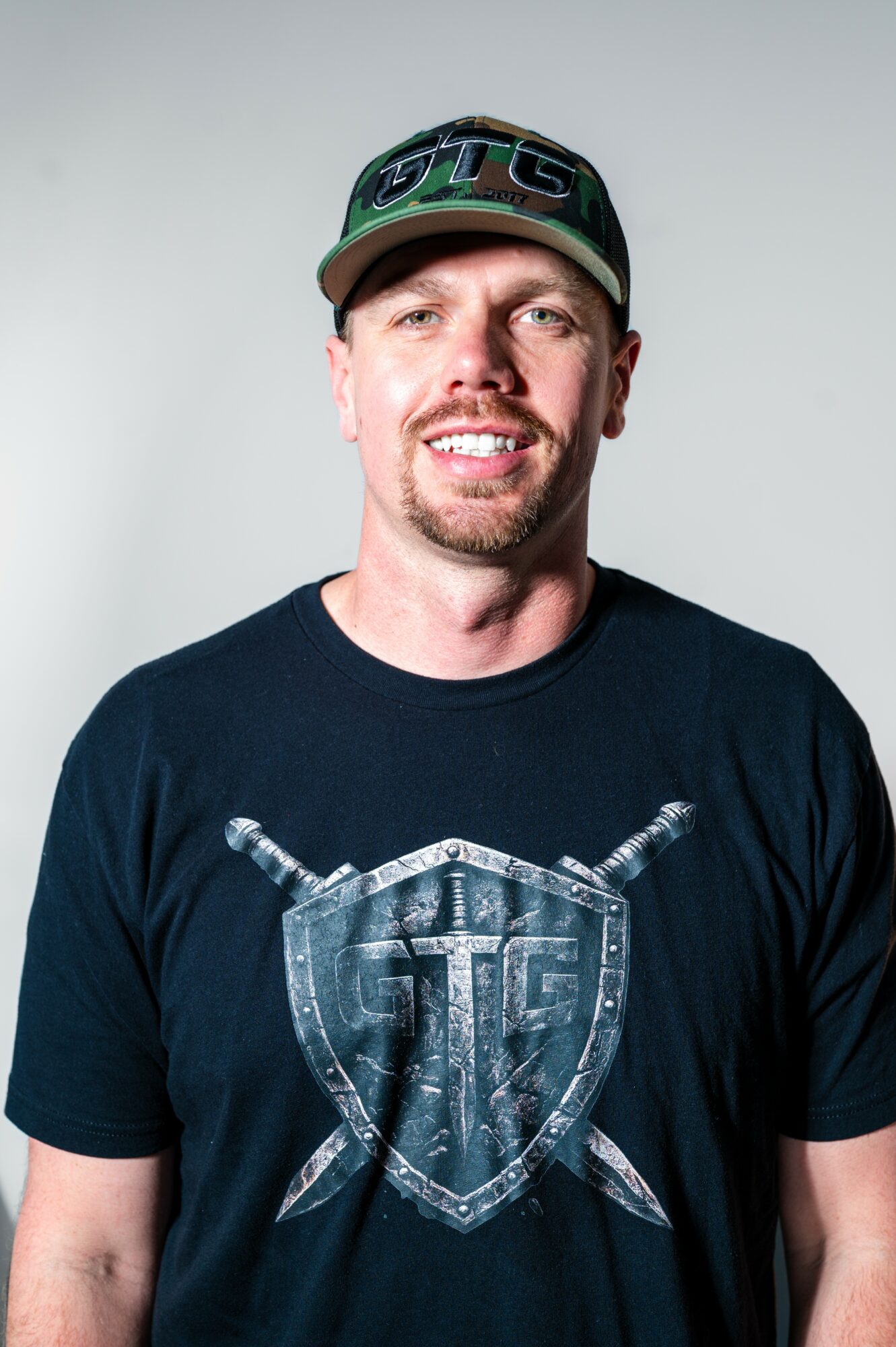 Smiling man wearing a black cap and T-shirt with a shield and crossed swords design, against a plain background.