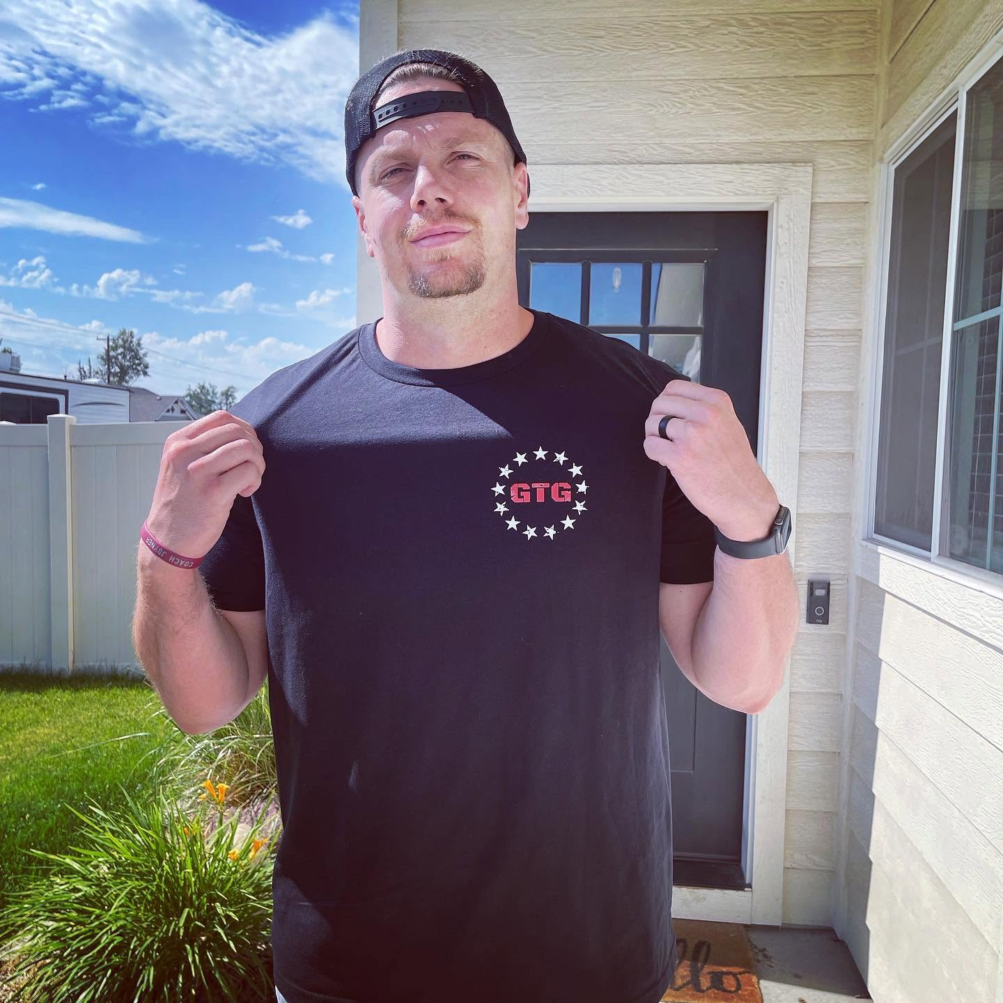 Young man with a cap, wearing a black T-shirt, standing outside near a house door, smiling, holding the shirt's collar.