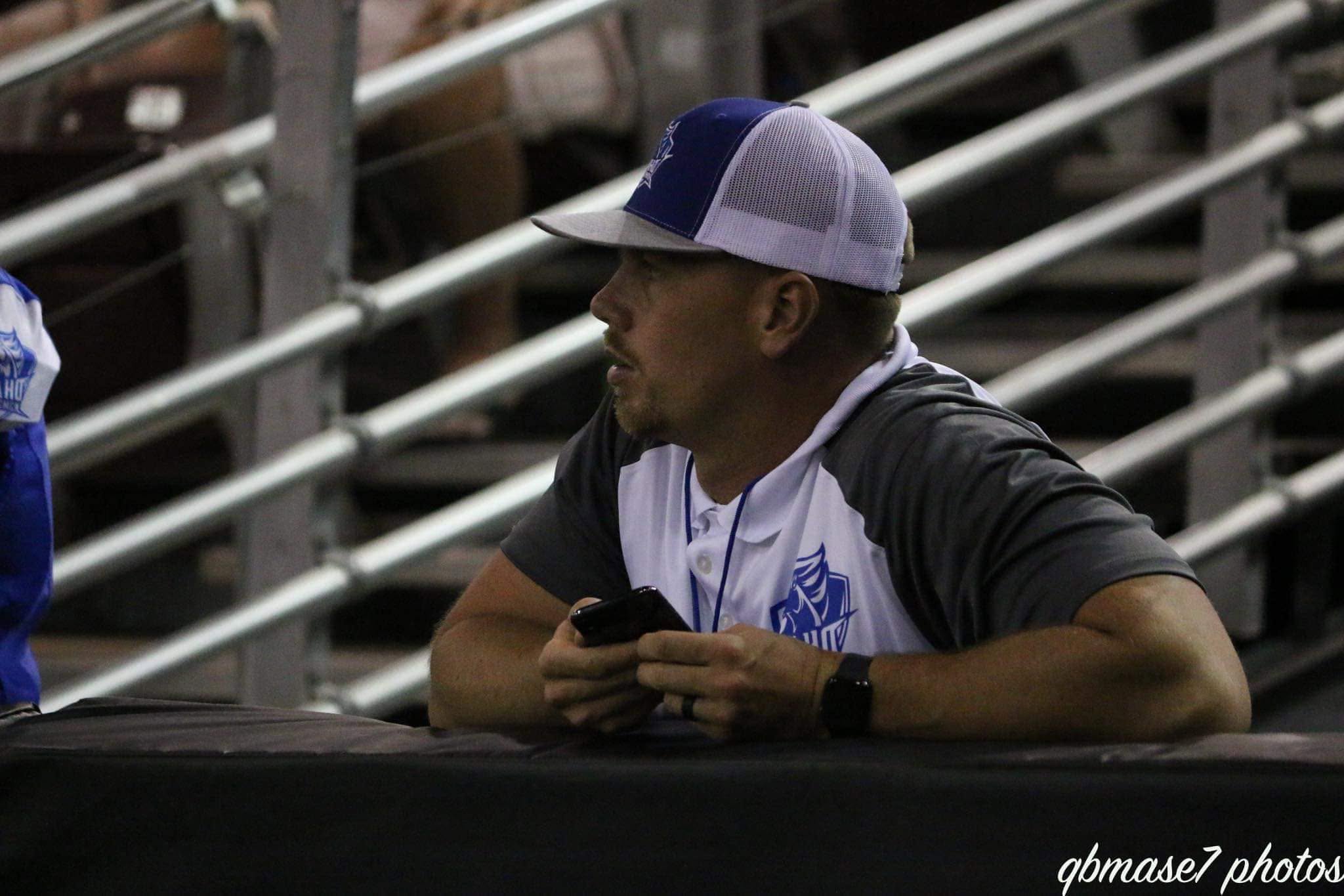Man wearing a baseball cap and casual clothing sitting on bleachers, holding a phone, with metal railings in the background.