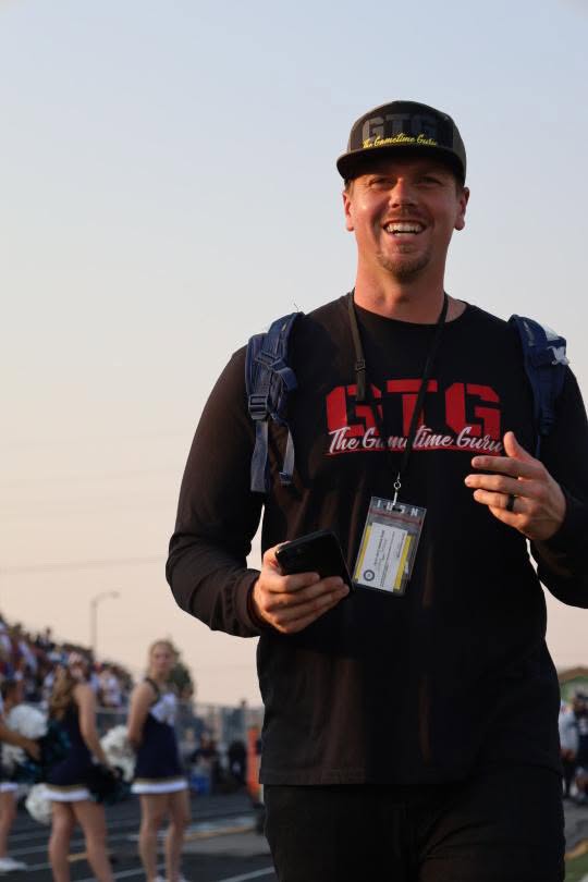 Young man smiling, wearing a black cap, black hoodie, and backpack, holding a phone and pointing with his finger outdoors.
