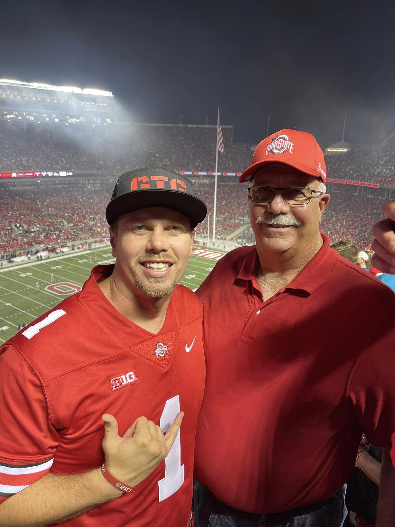 Two men in red Ohio State and football gear at a stadium, smiling, night setting, stadium lights in background.