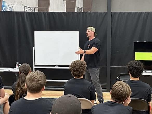 Man standing in front of a whiteboard, speaking to a group of seated people in a room with black walls.