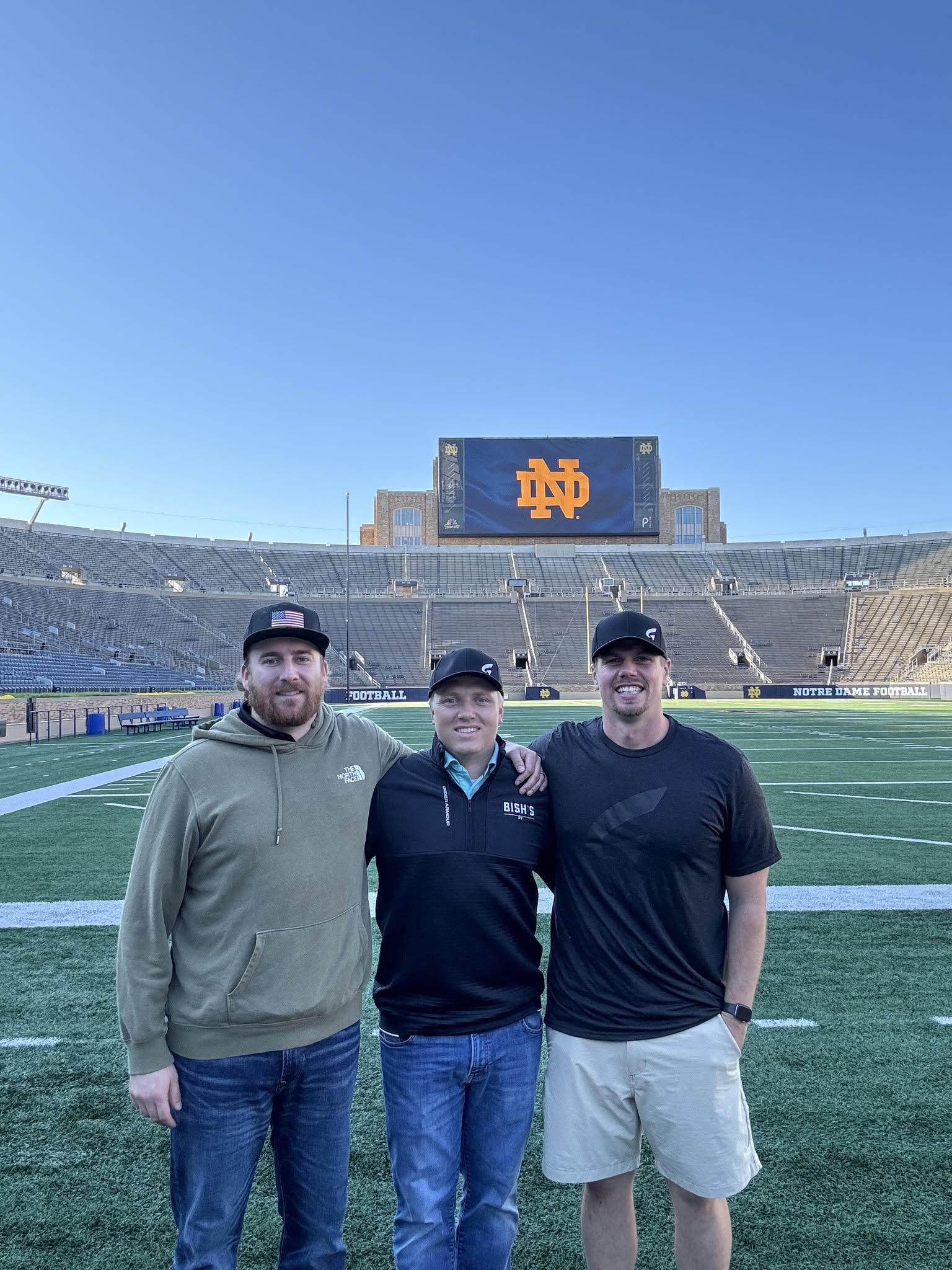 Three men standing on a football field with empty stadium seats and a large screen displaying the Notre Dame logo in the background.