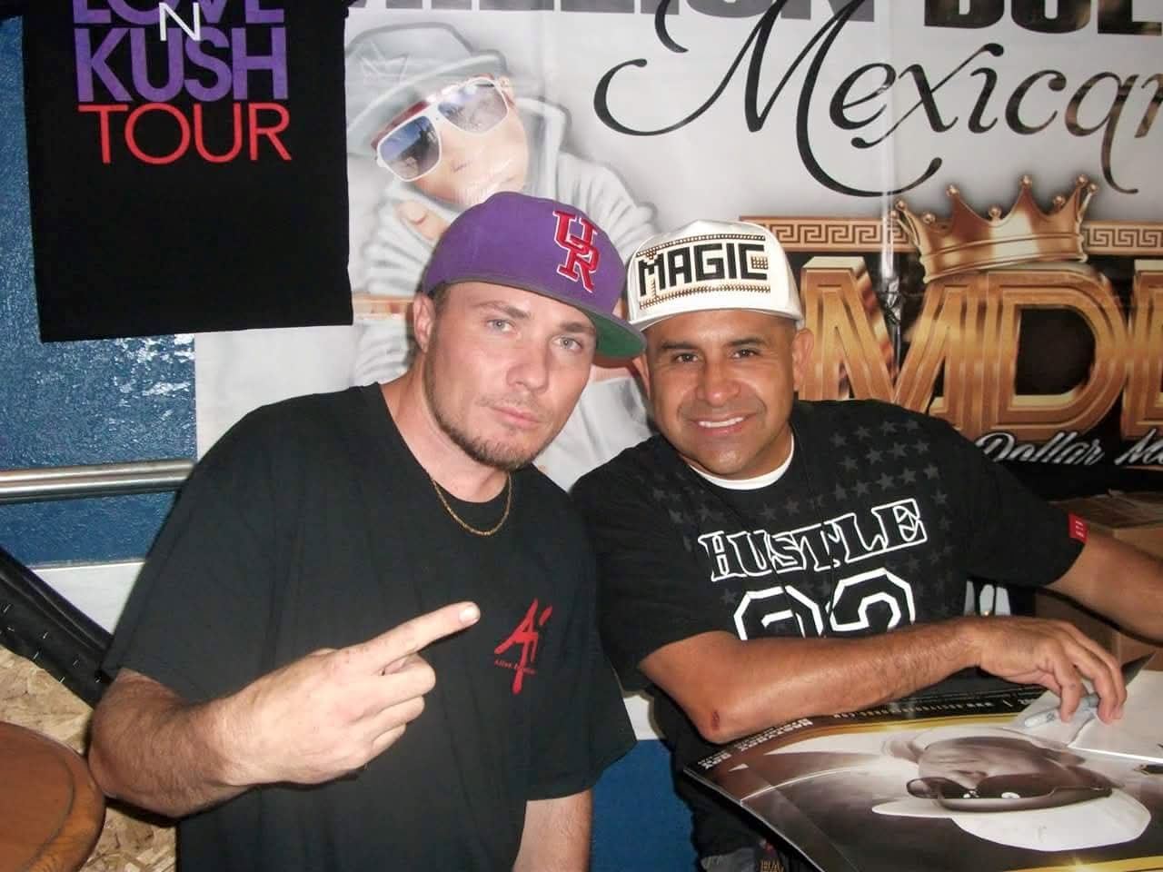 Two men smiling, one pointing, sitting at a table with promotional posters in the background.