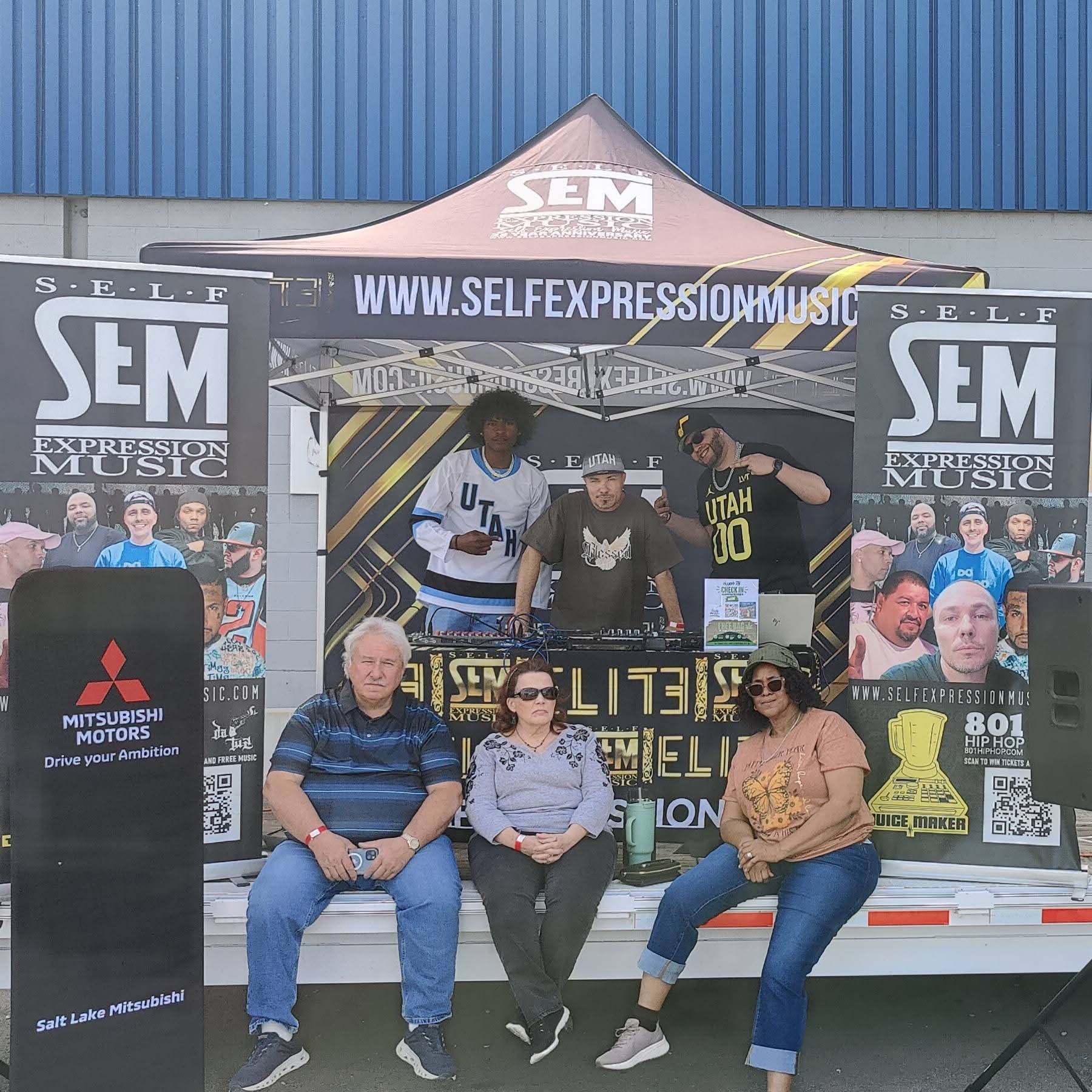 Group of four people sitting and standing in front of a music event booth with banners and a tent, outdoors.