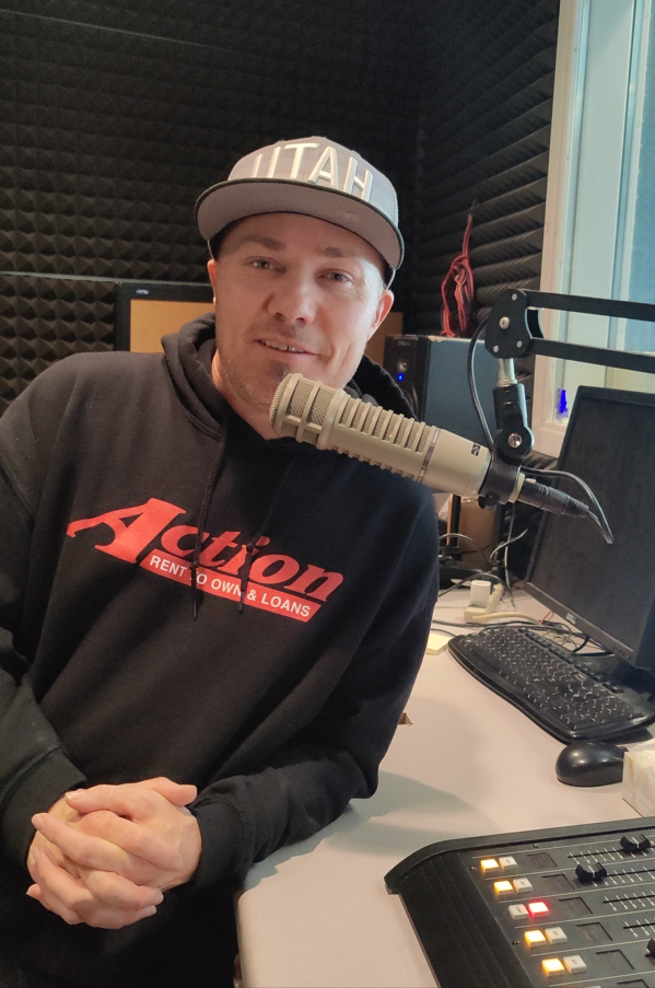 Person wearing a cap and hoodie, sitting in a recording studio with a microphone and audio equipment.
