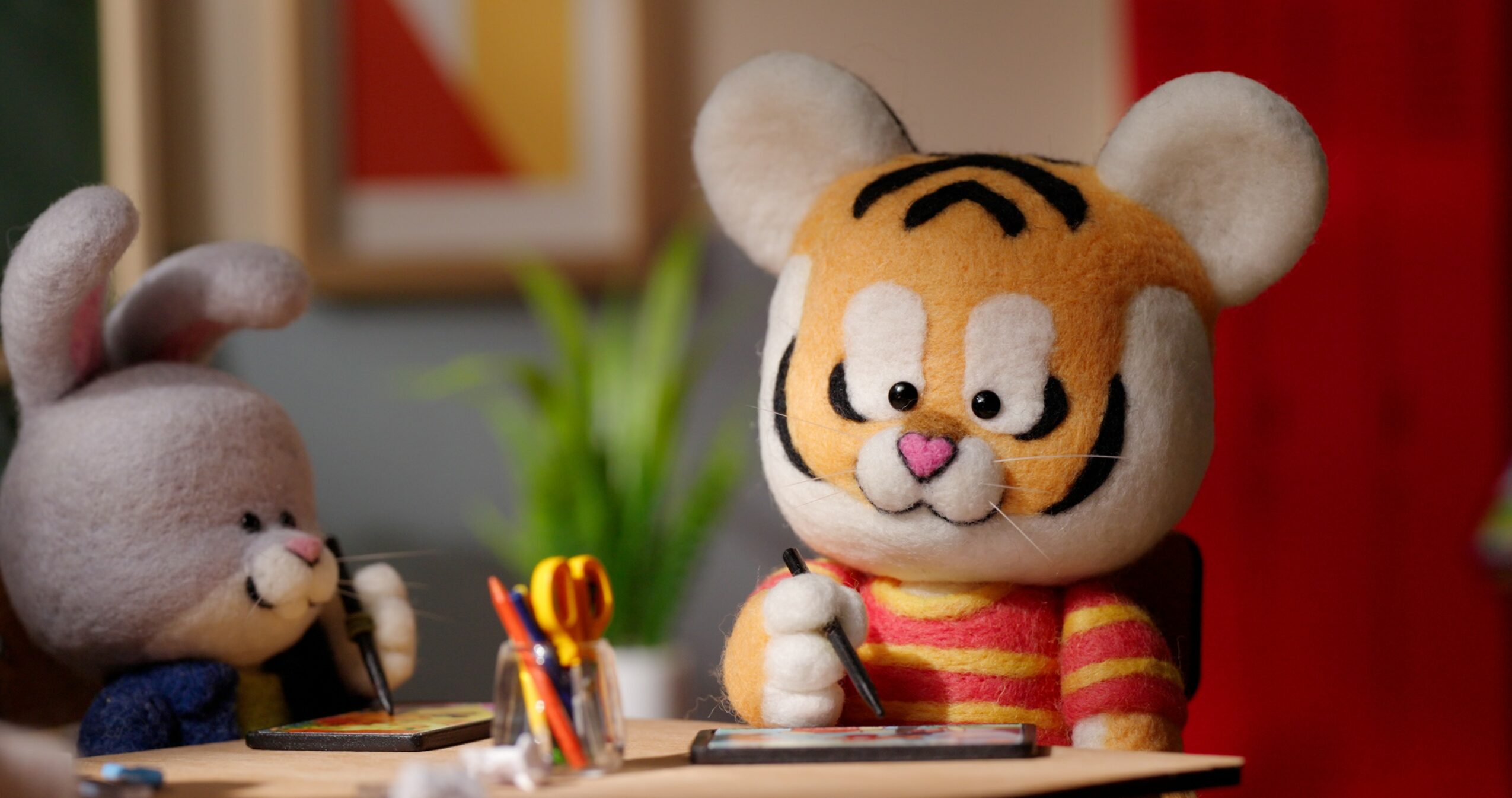 Two plush animal characters, a rabbit and a tiger, sit at a desk with stationery, in a colorful room.