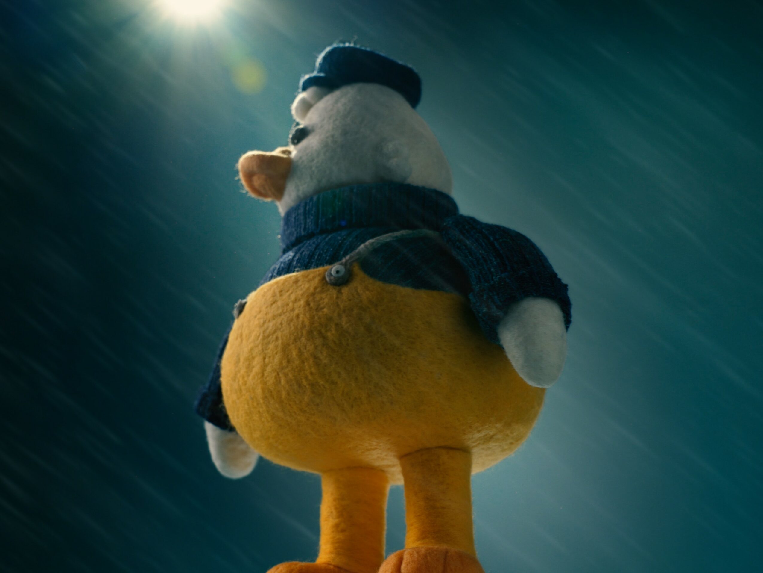 A plush toy character with a large yellow bottom, wearing a blue hat and scarf, facing away in a dark background.