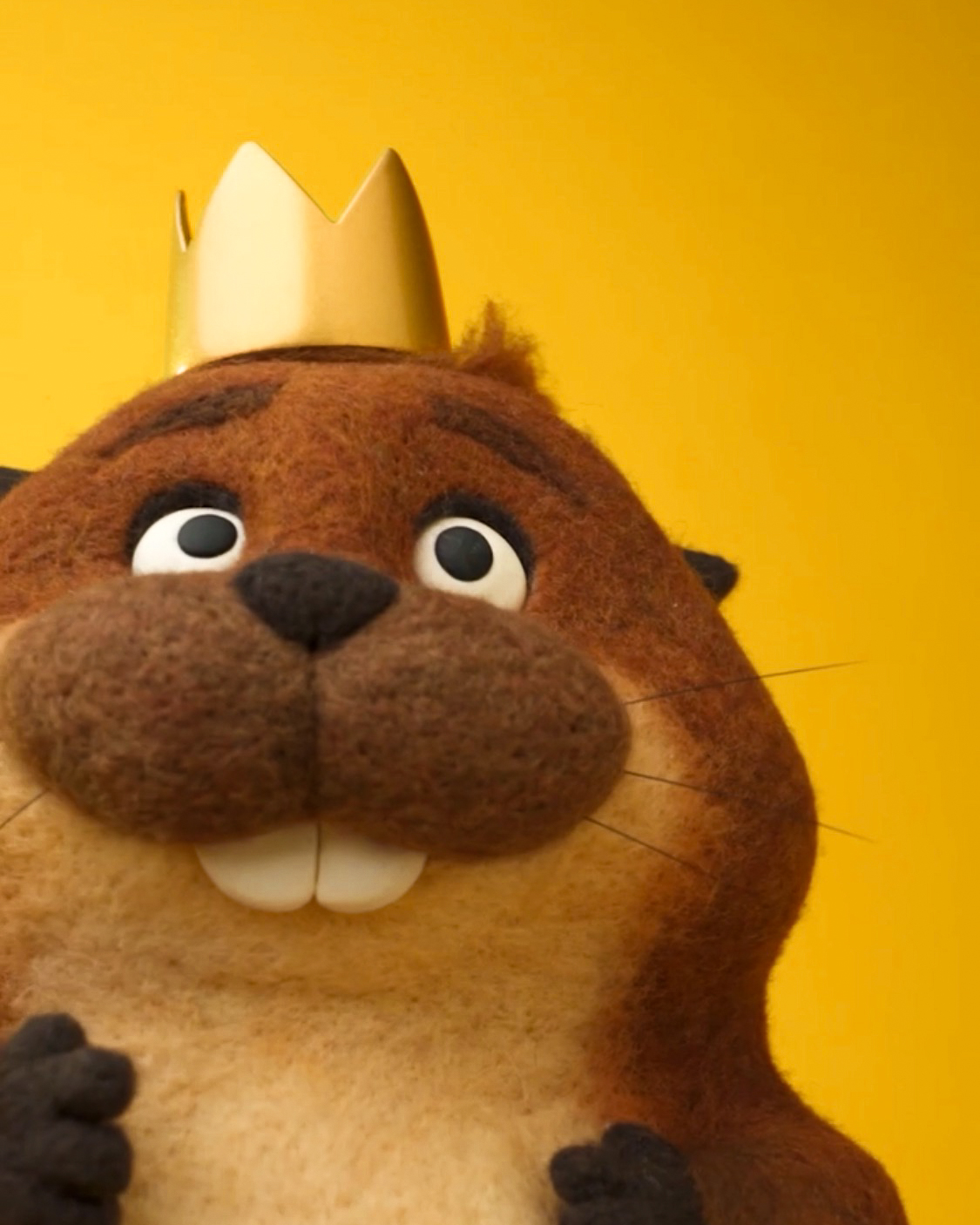 Close-up of a puppet otter with a yellow background, wearing a paper crown, showing teeth and wide eyes.