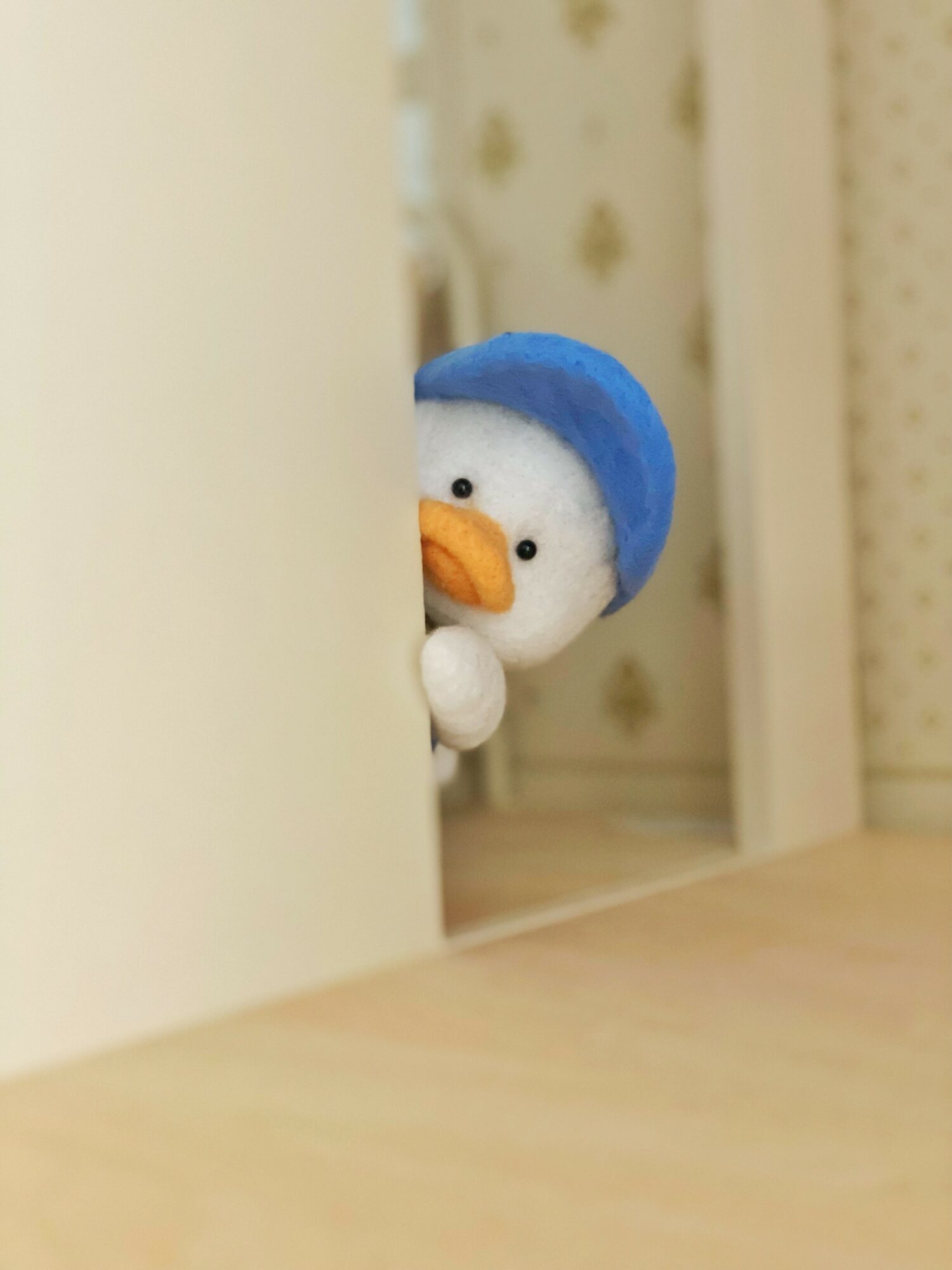 A plush penguin toy peeks from behind a piece of furniture, wearing a blue hat and holding a white object.