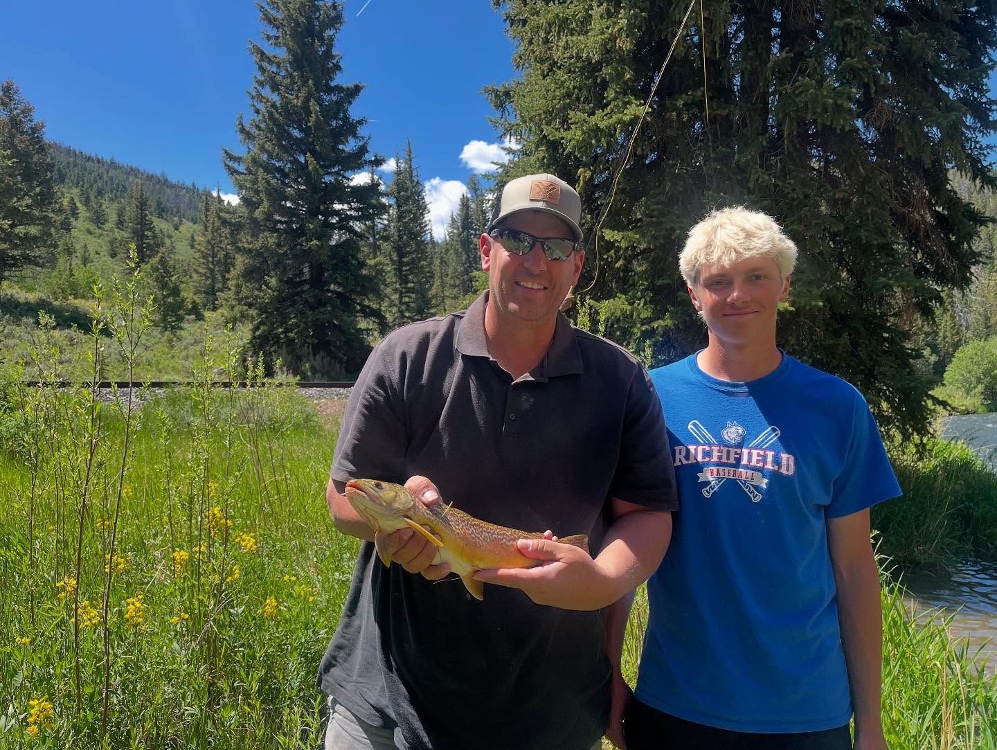 Two people outdoors in a grassy area with trees, one holding a fish, both smiling, sunny day.