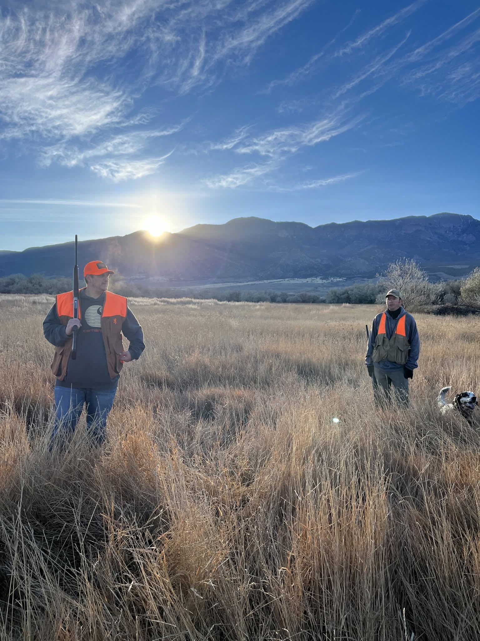 Two people wearing orange vests and hats walk through a grassy field with mountains and a setting sun in the background.