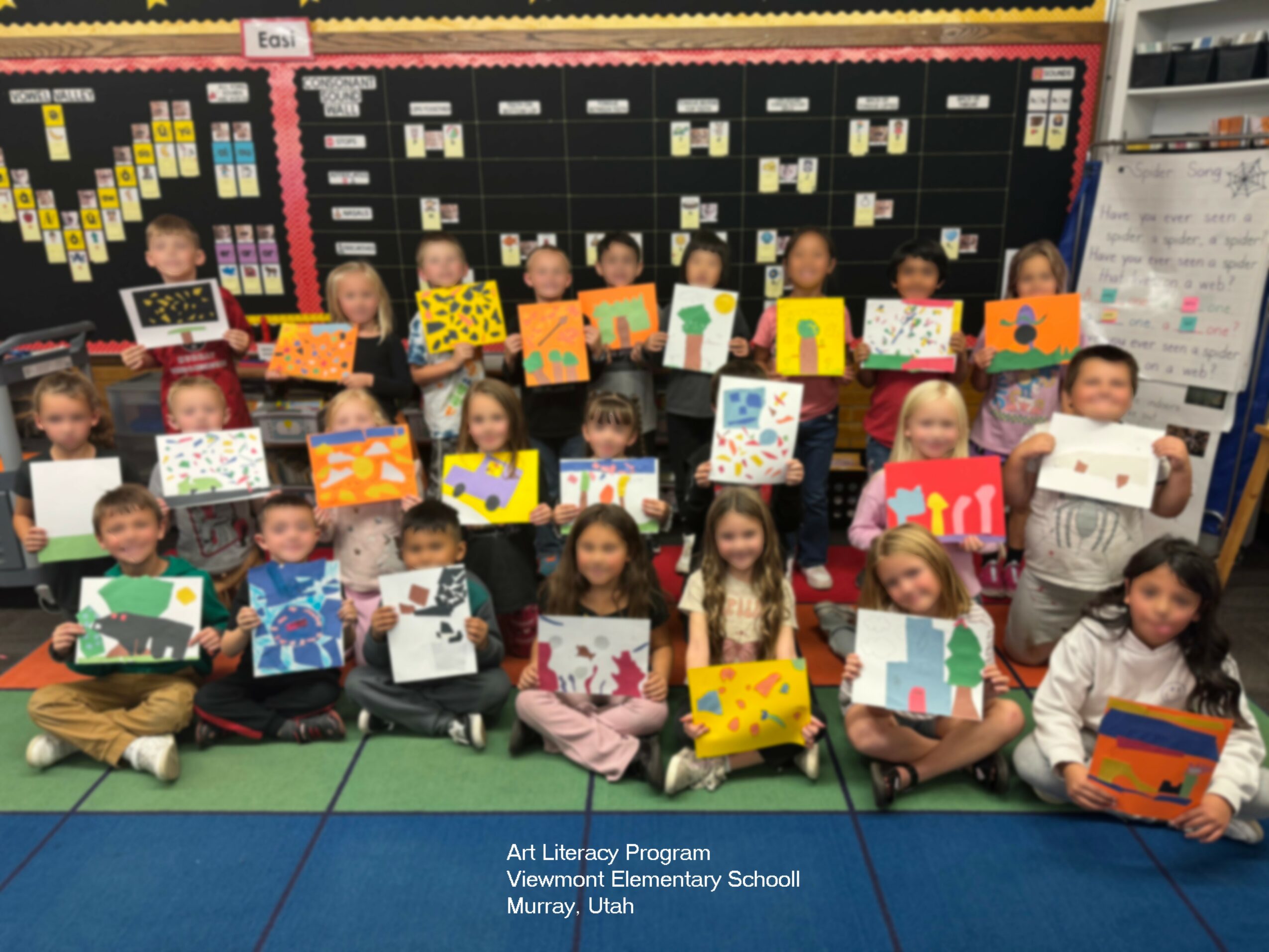 Group of children holding colorful artwork in classroom with educational decorations and a whiteboard in background.
