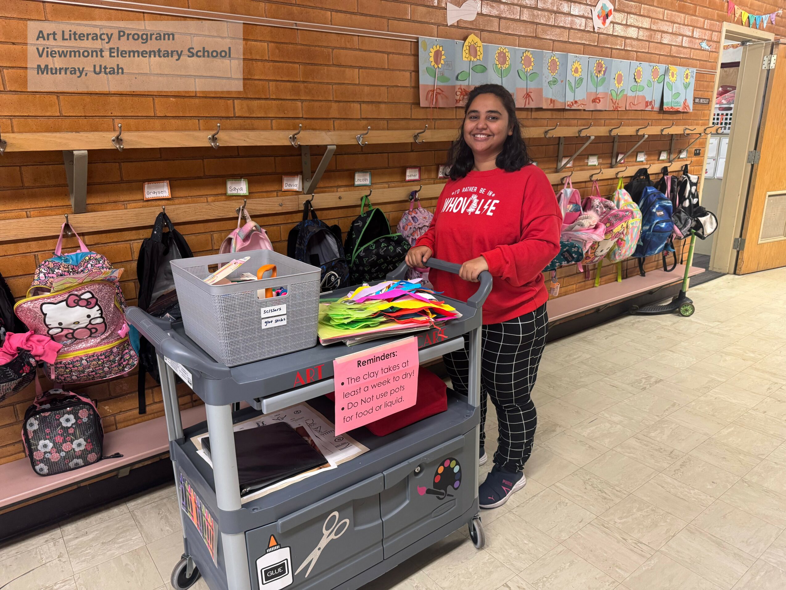 Girl standing next to a table with school supplies, smiling, in a school hallway with backpacks and a bulletin board.