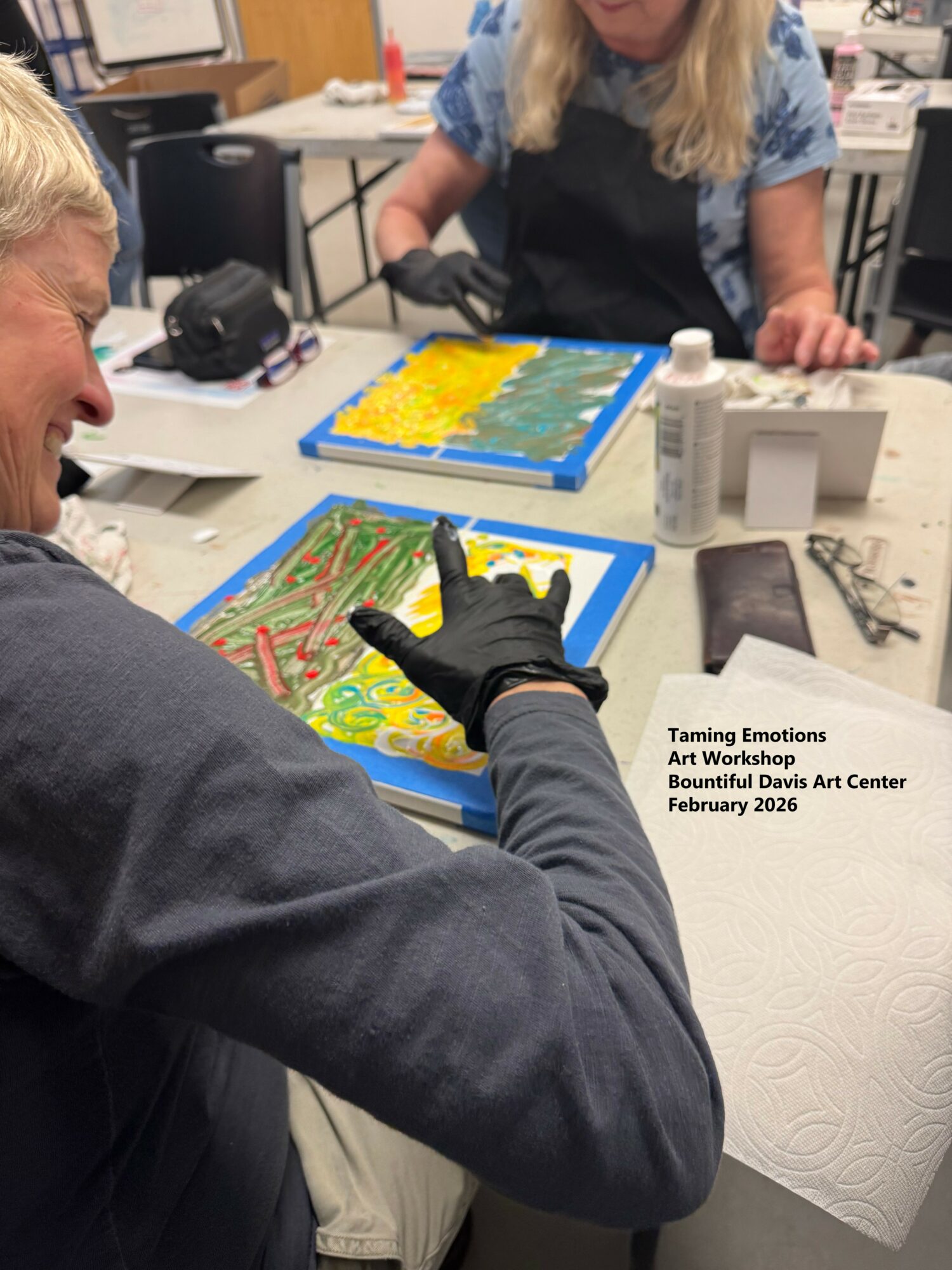 Two women working on colorful abstract art projects at a table, with art supplies and a paper towel nearby.