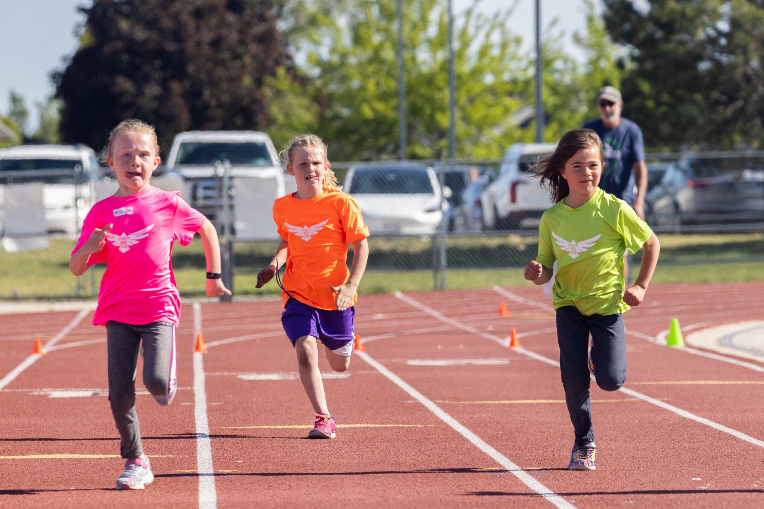 Three children running on a track with a person watching in the background, trees, and parked cars, sunny day.