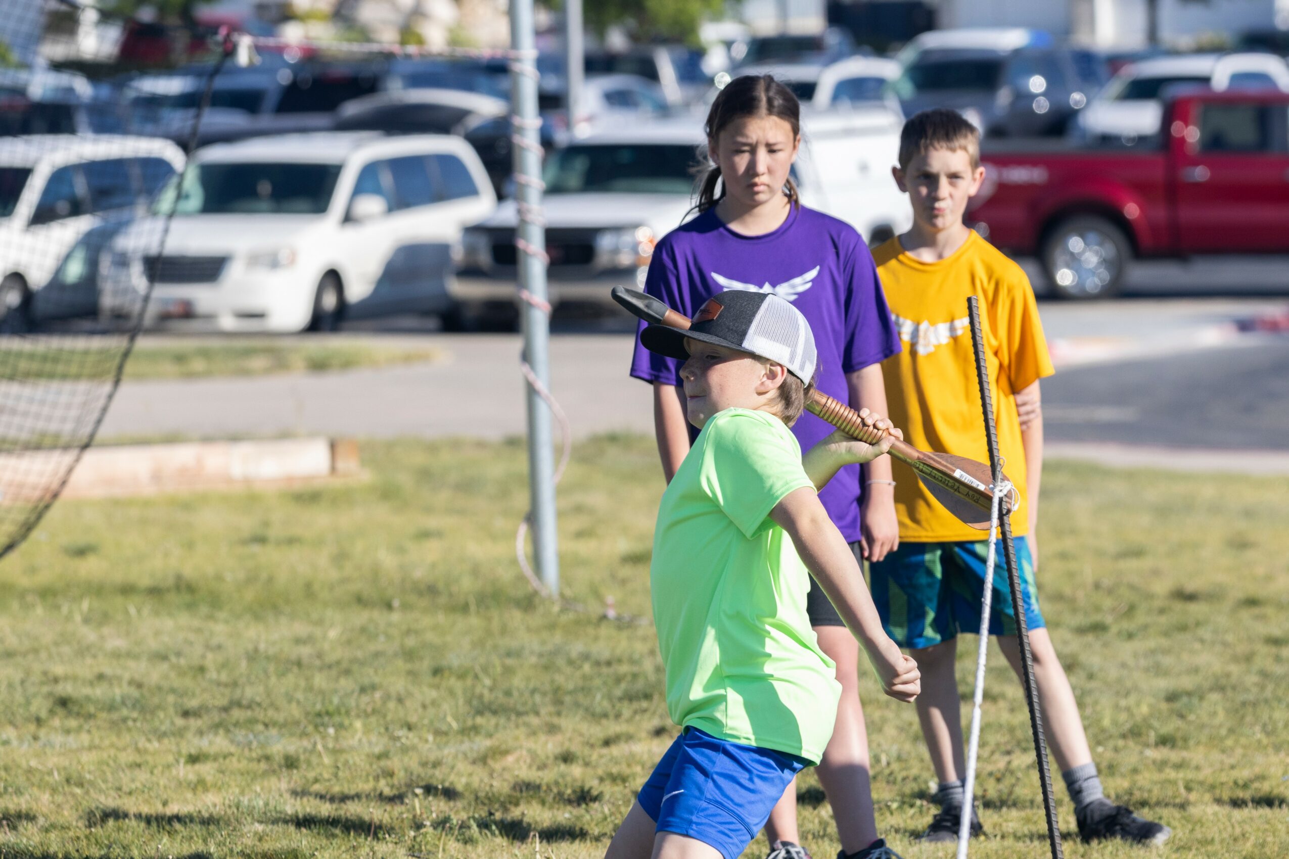Three children playing with a stick outdoors on grass, with parked cars in the background.