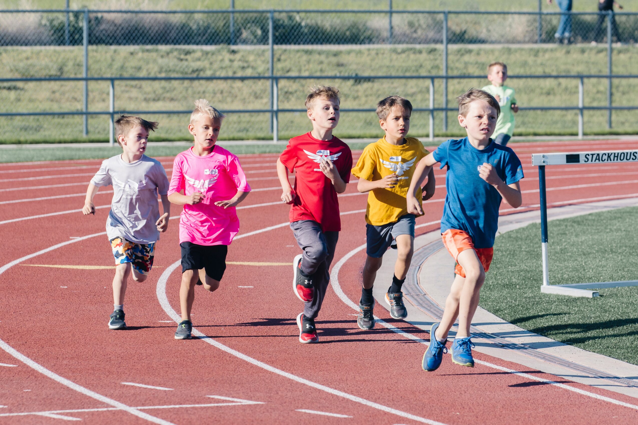 Six children running on a track during a race, wearing colorful athletic clothing, with a grassy field and fence in background.