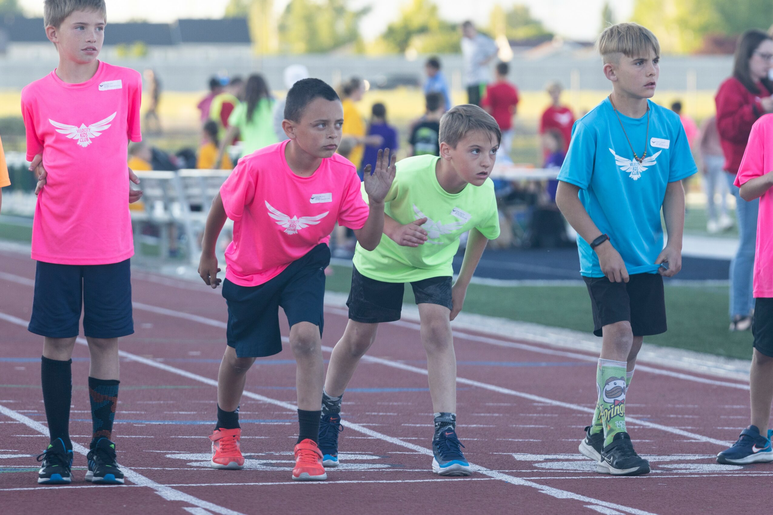 Four children in colorful sports shirts and shorts preparing to race on a track, with other children in the background.