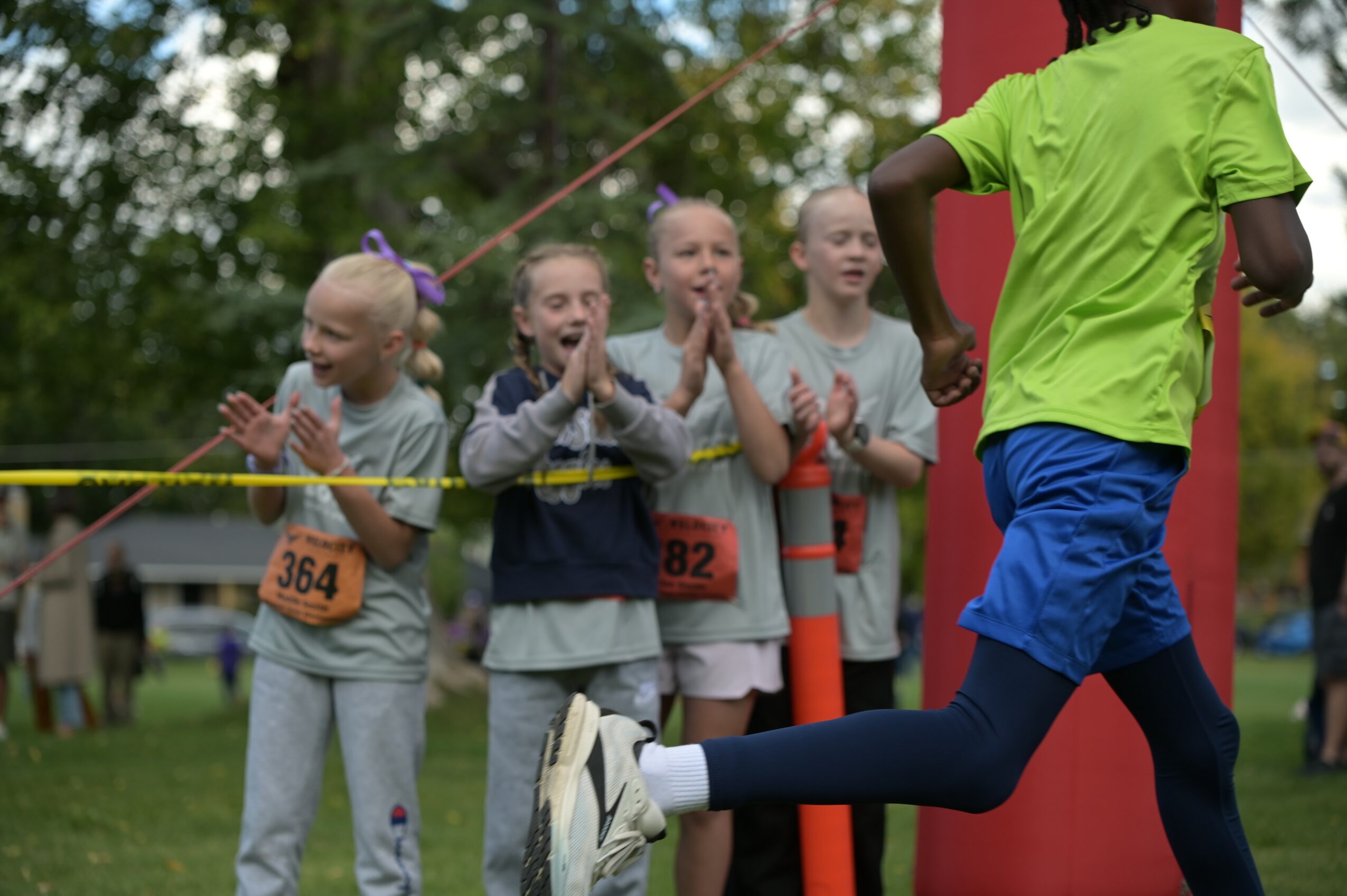 Children watch a runner in a green shirt and blue shorts during a race outdoors, with trees in the background.