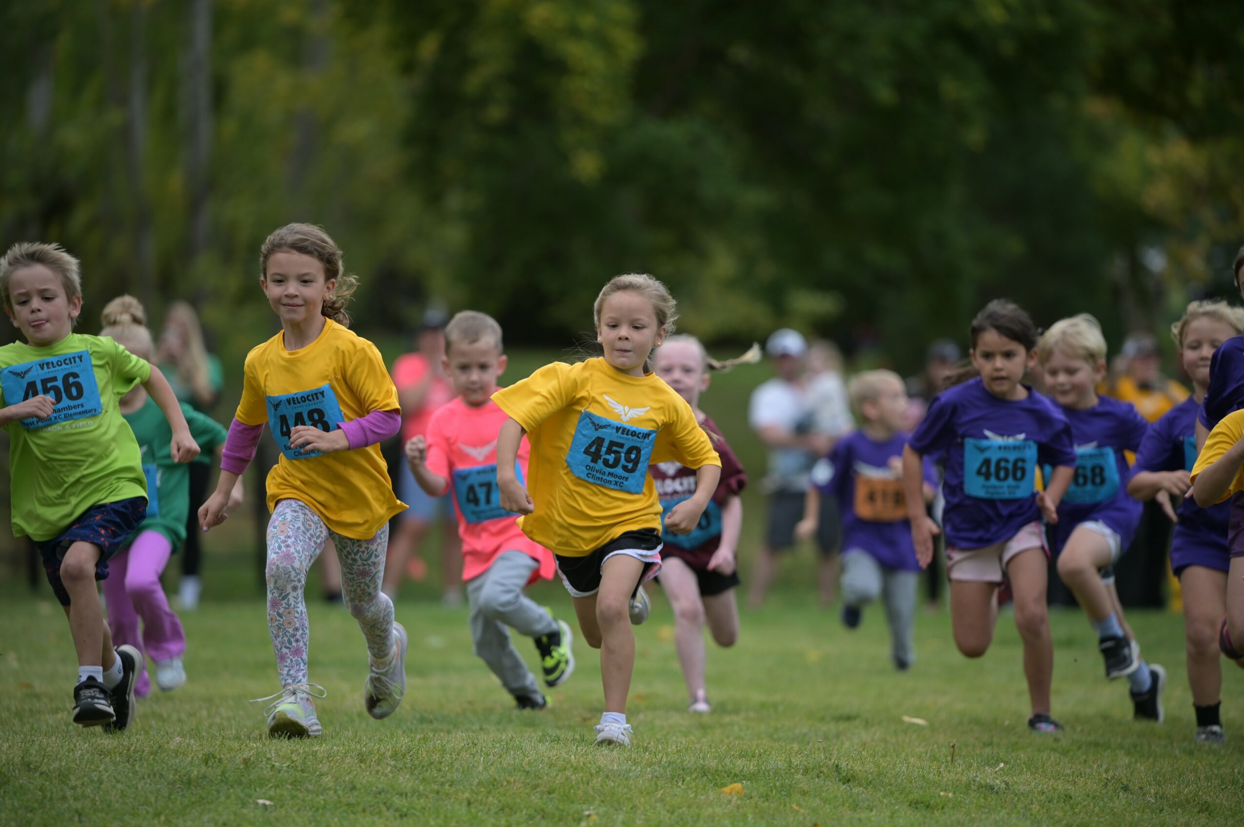 Children running in a race on a grassy field with trees in the background, wearing colorful athletic clothing and numbered bibs.