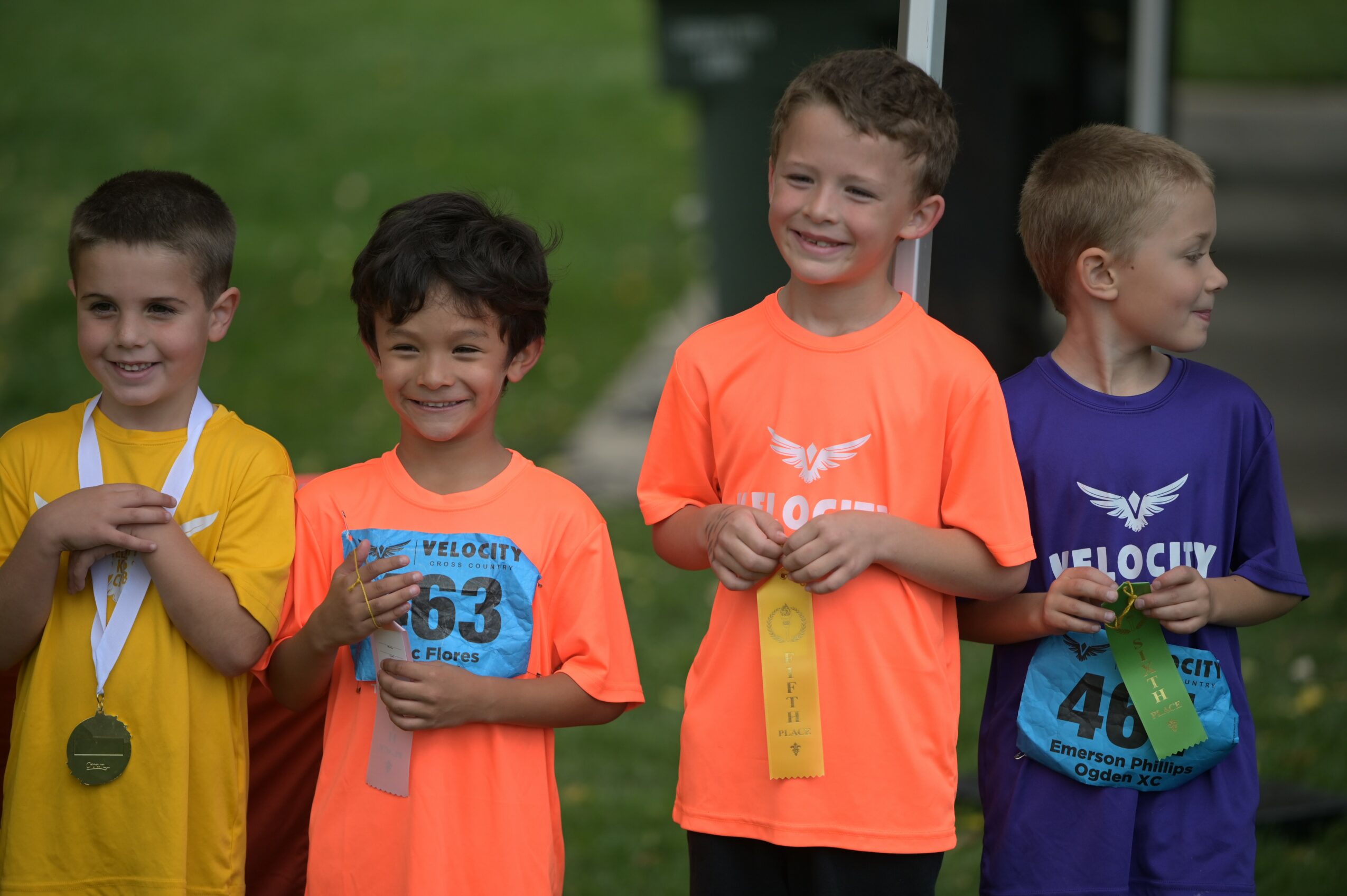 Four children stand outdoors, smiling, wearing colorful athletic shirts and medals, holding ribbons, with a grassy background.