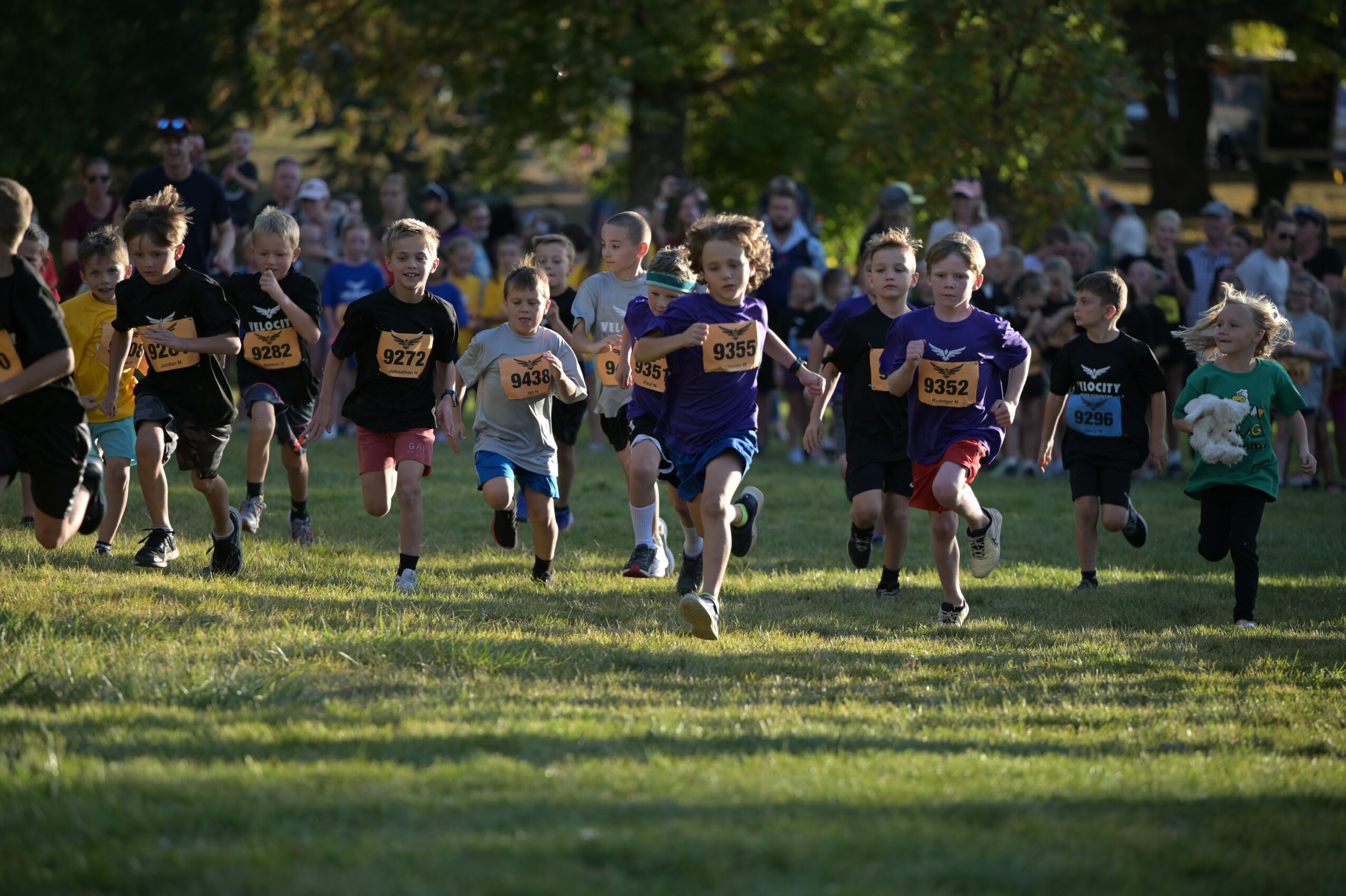 Children running in a race outdoors on grass with trees in the background, some wearing numbered bibs.