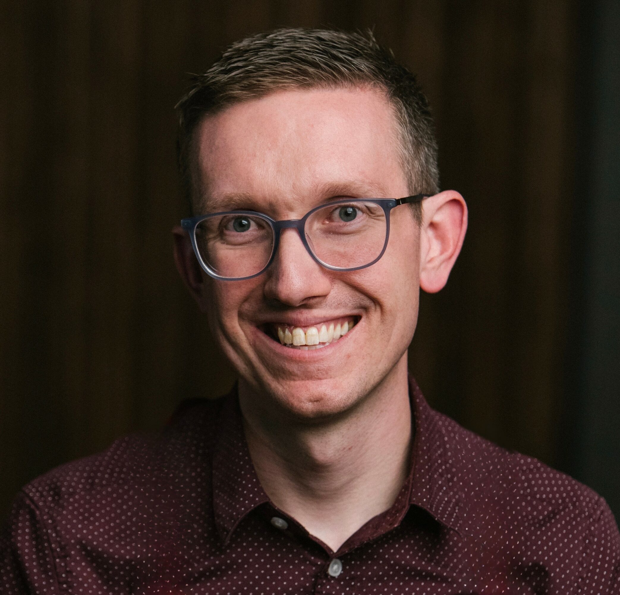 Smiling man with glasses and short hair, wearing a maroon shirt, against a dark background.
