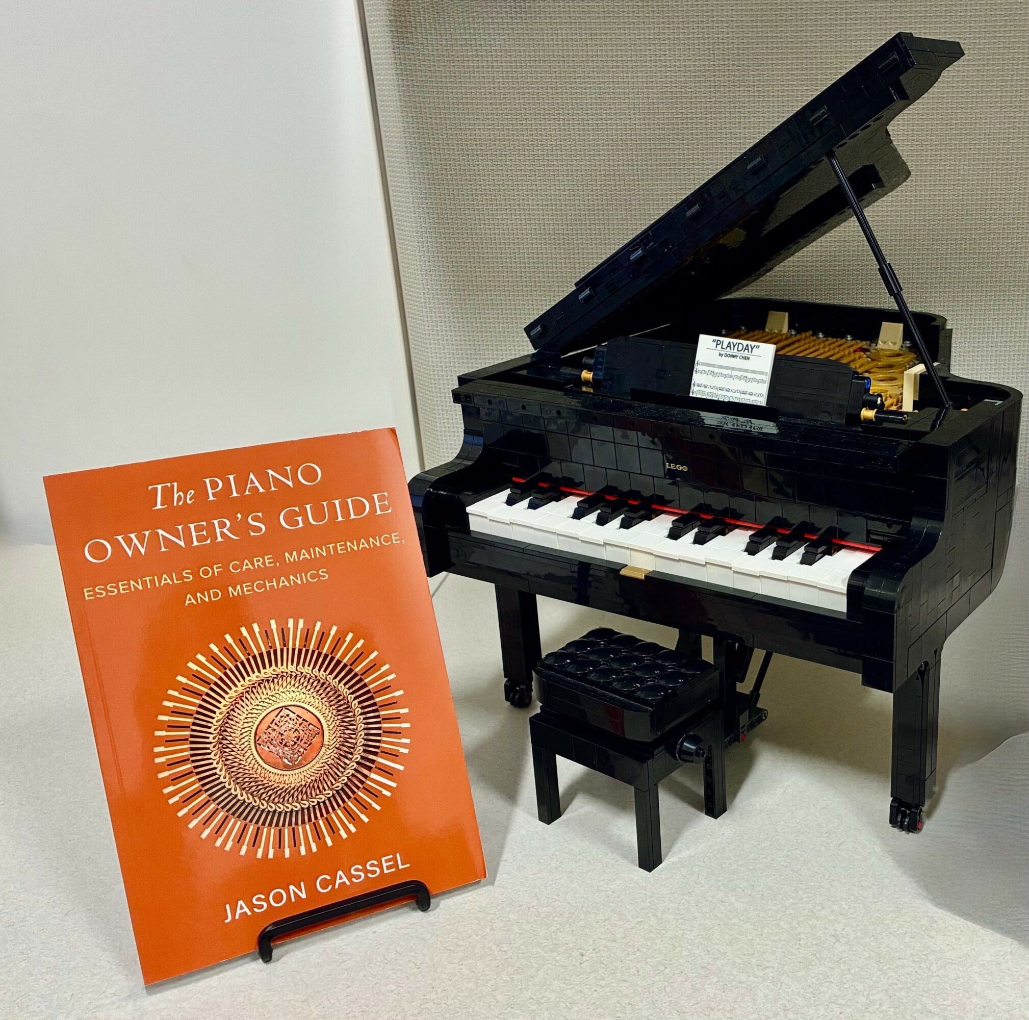 Small black grand piano with open lid, white and black keys, and a stool in front, next to a red book titled 'The Piano Owner's Guide'.