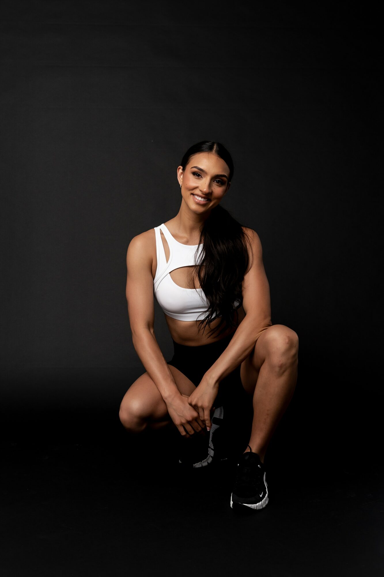 Young woman with long dark hair in athletic wear, smiling, kneeling on a black background.