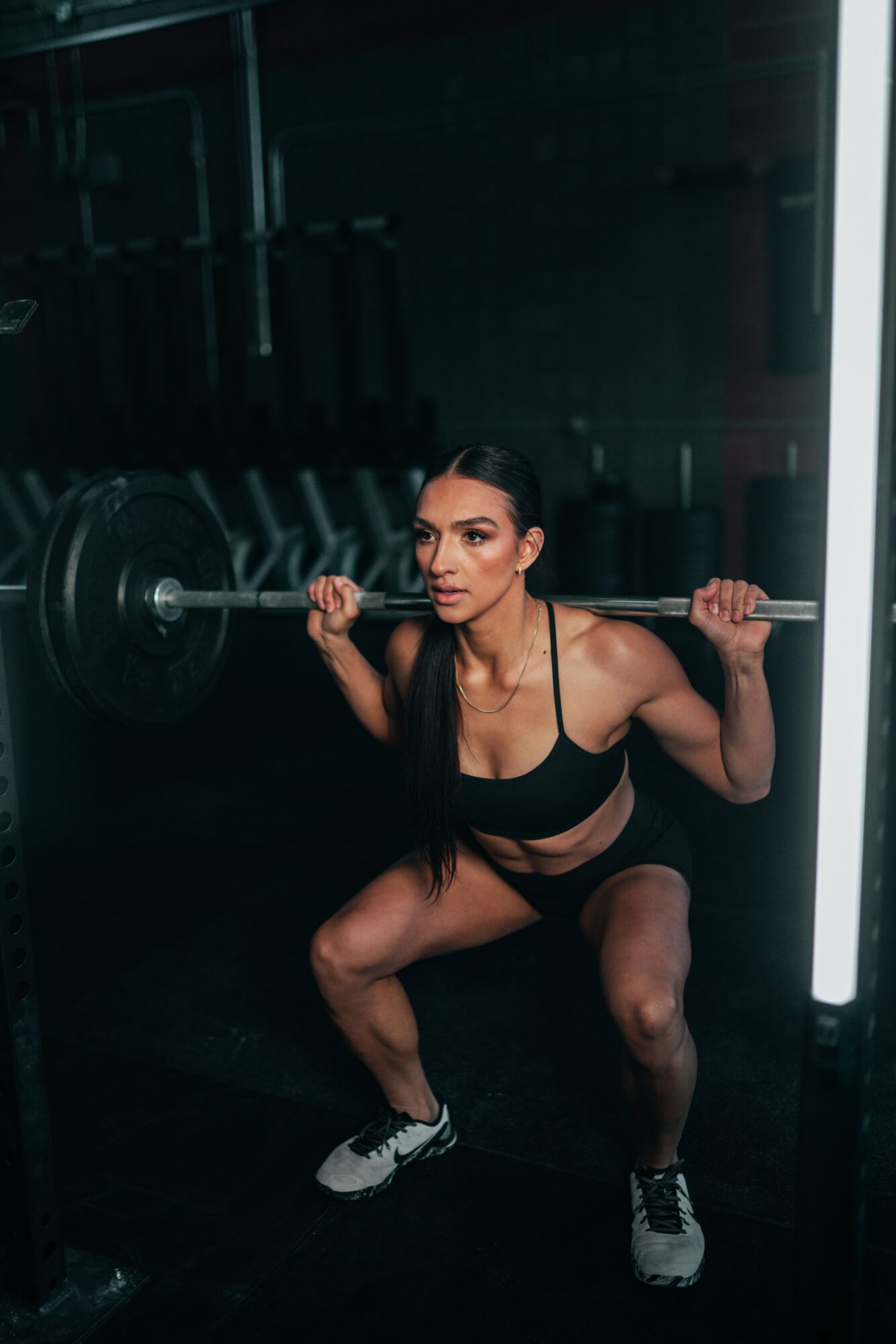Woman in gym squatting with barbell on shoulders, dark background, illuminated vertical light, wearing sports bra and shorts.