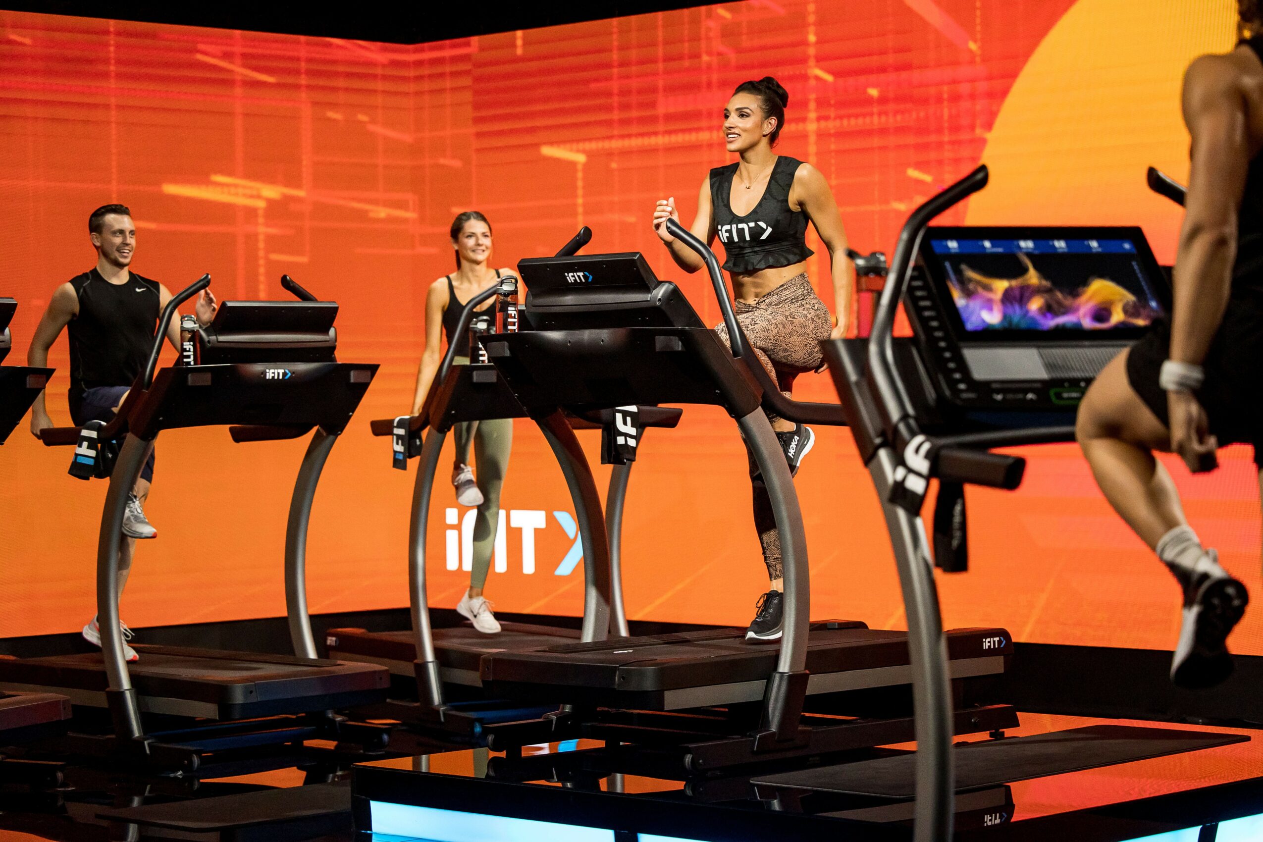 People exercising on treadmills in a gym with a bright orange background, some wearing workout clothes and glasses.