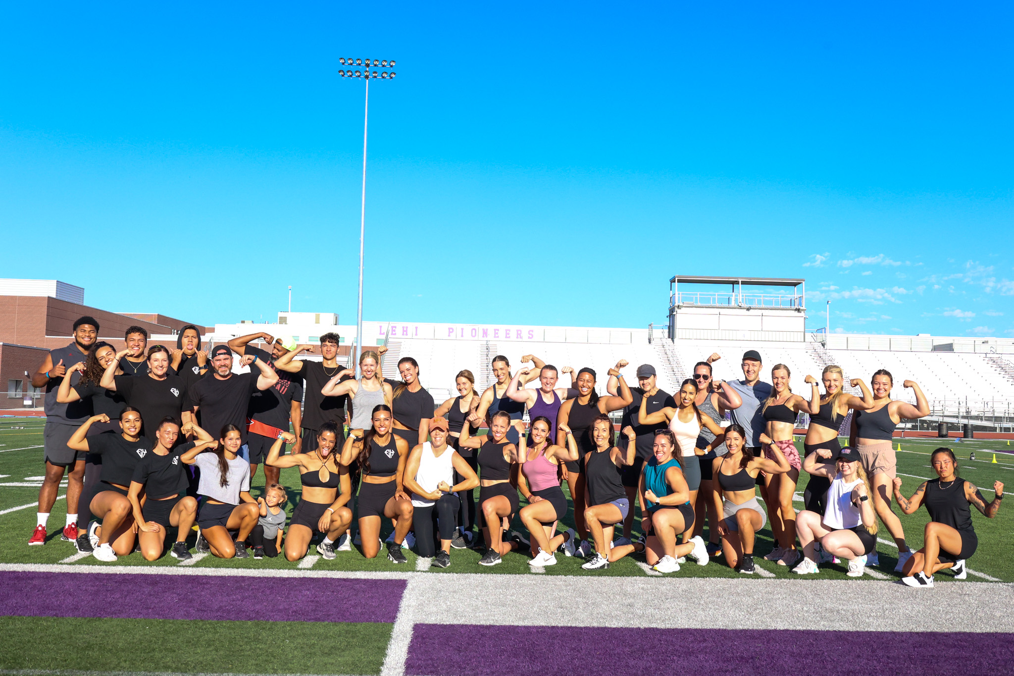 Group of people on a sports field, some standing and some kneeling, with a clear blue sky in the background.