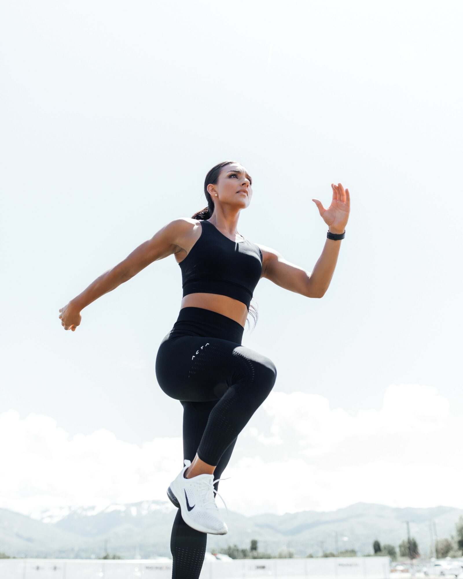 Woman jumping outdoors with arms raised, wearing athletic clothing and sneakers, against a cloudy sky background.