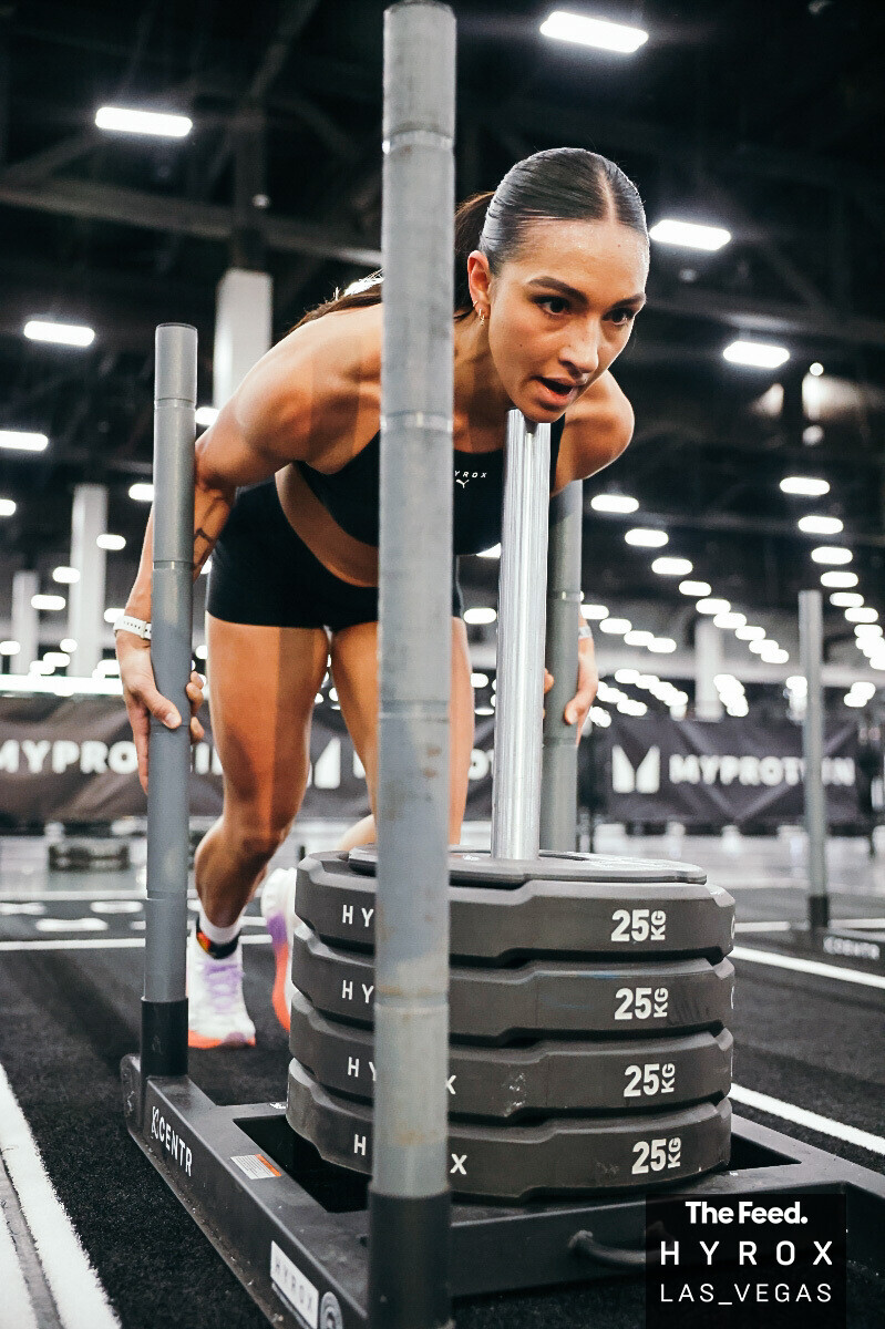 Female athlete in a gym, pushing weighted sled, focused expression, gym equipment and bright lights in background.