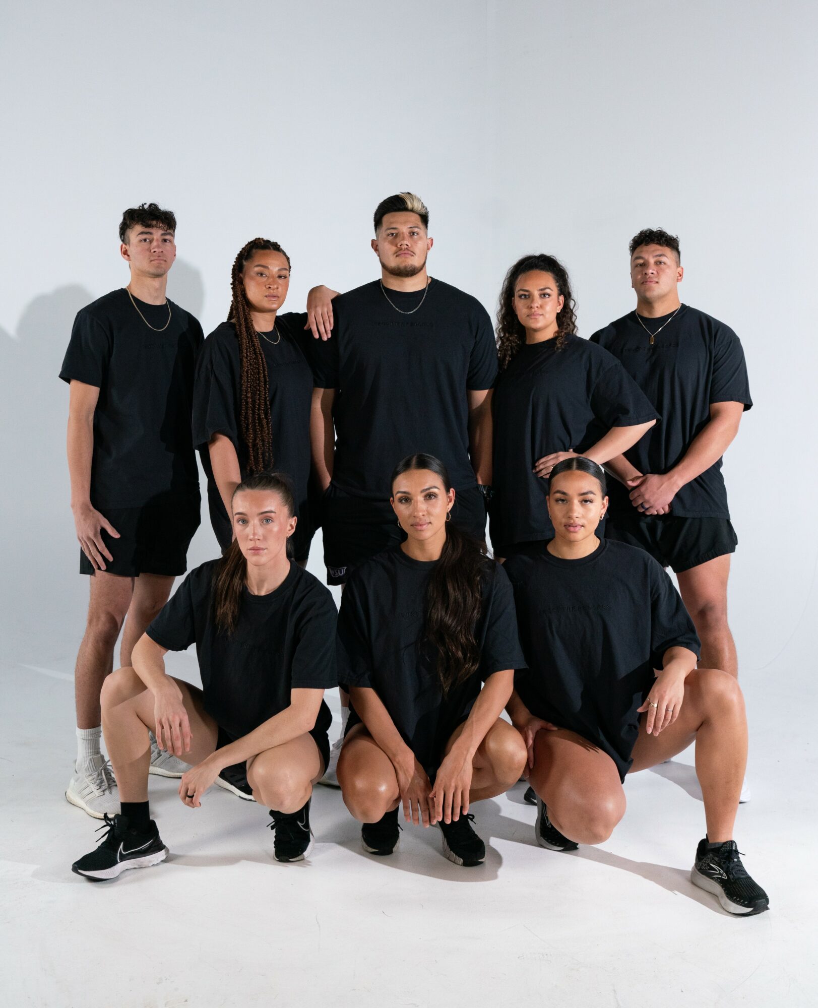 Group of seven young people in black athletic clothing, standing and kneeling against a plain white background.