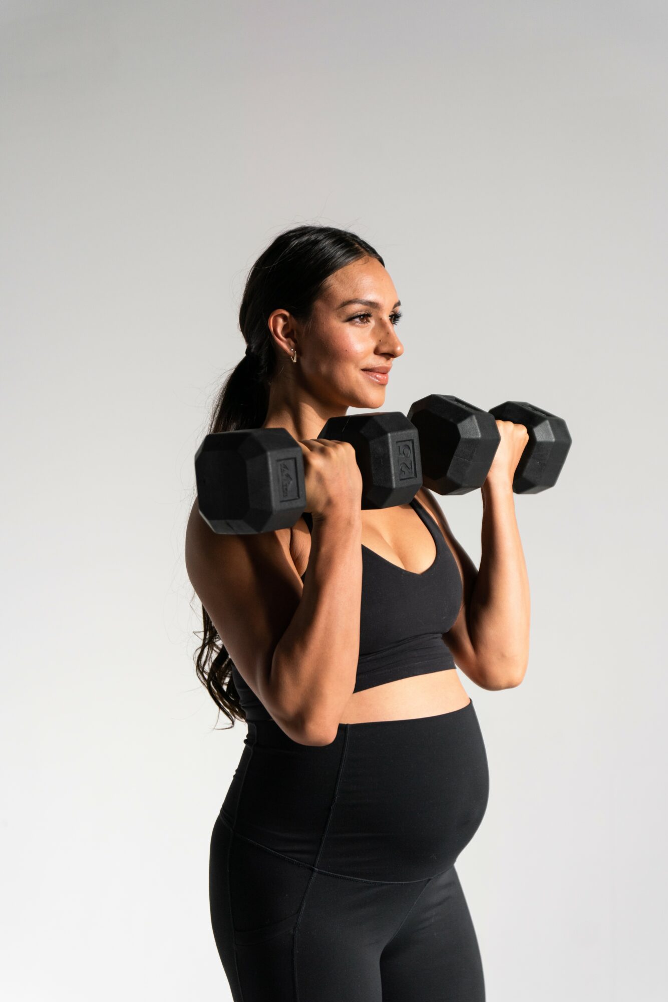 Pregnant woman lifting dumbbells with both hands, standing sideways, wearing black workout clothes, smiling, against a plain background.