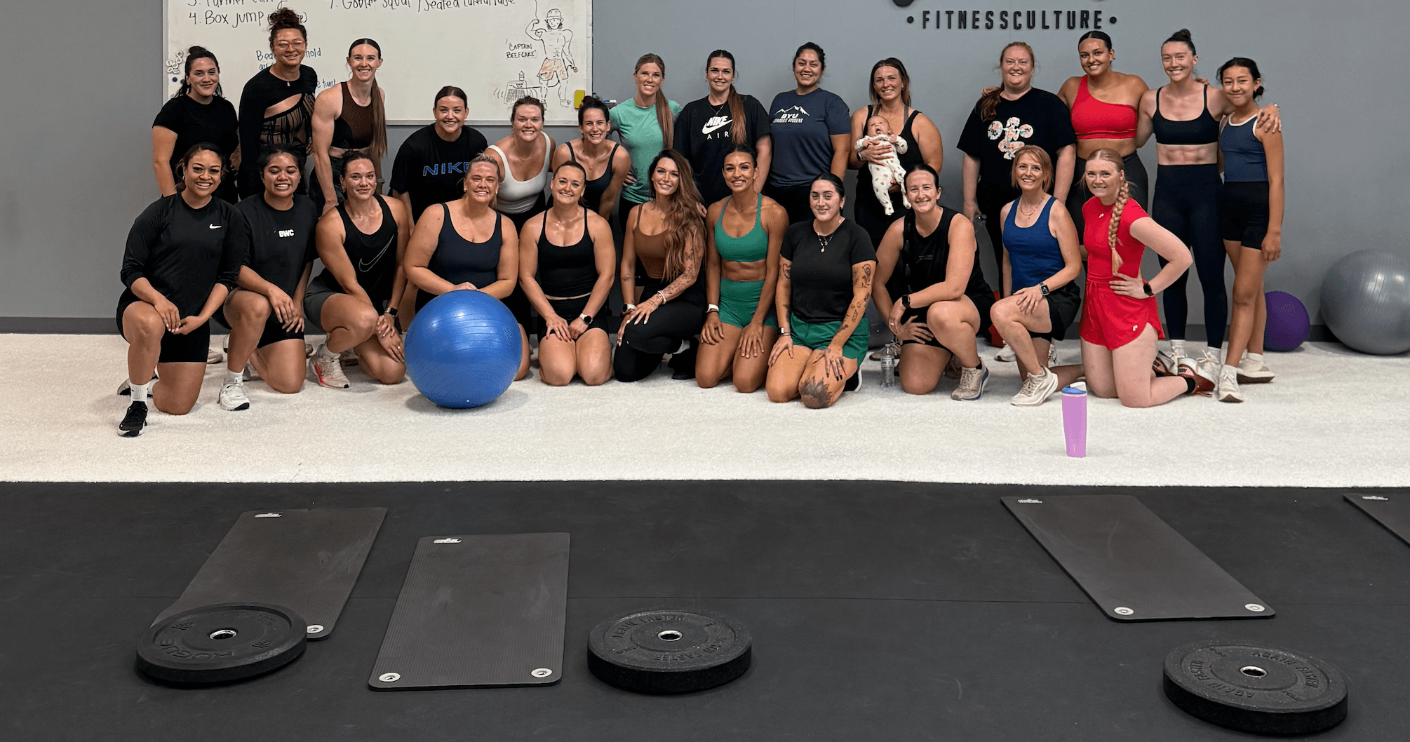 Group of people in workout attire posing indoors with exercise mats and fitness equipment.