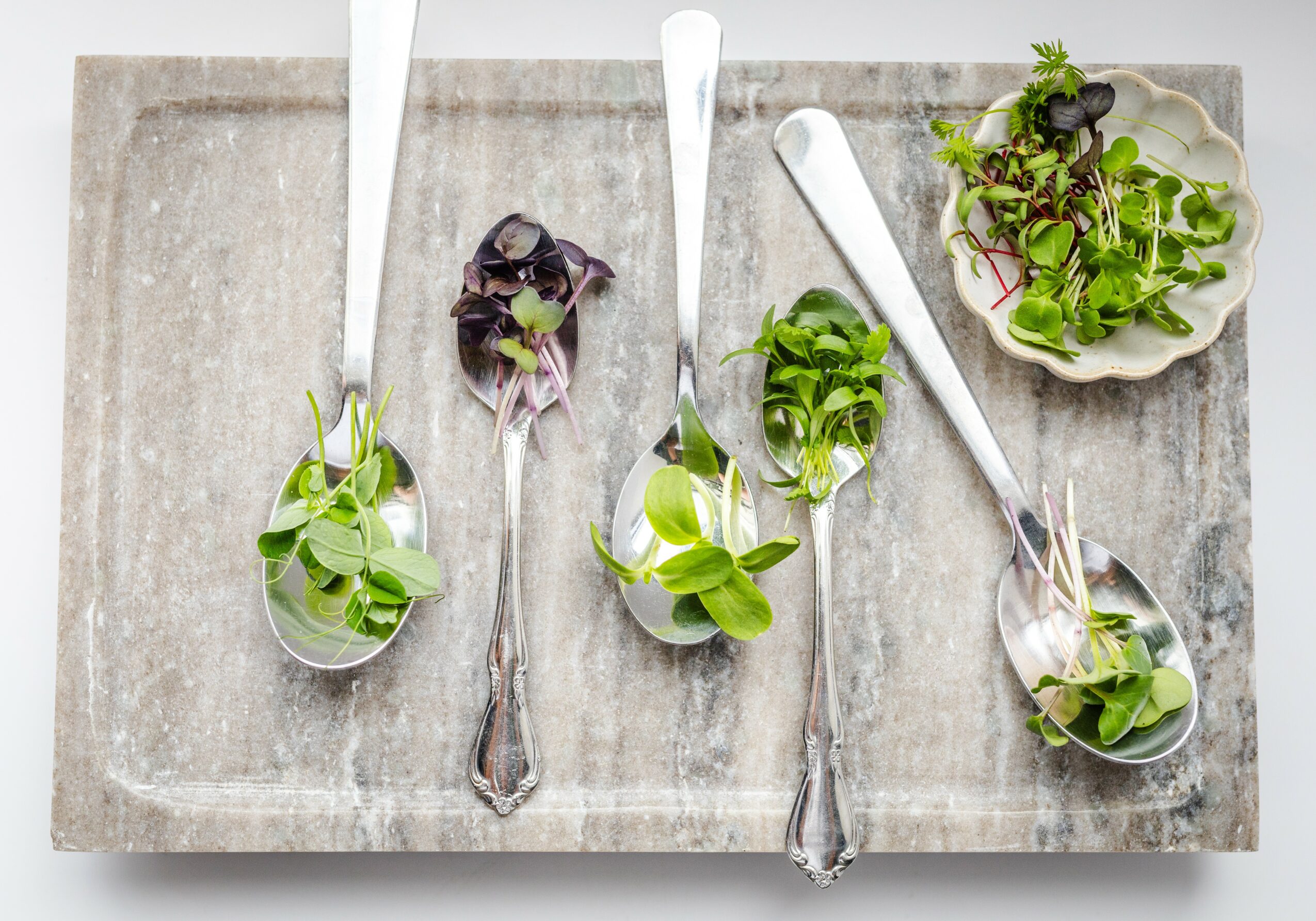 Four spoons with different types of fresh green herbs on a wooden tray, with a small bowl of herbs in the top right corner.