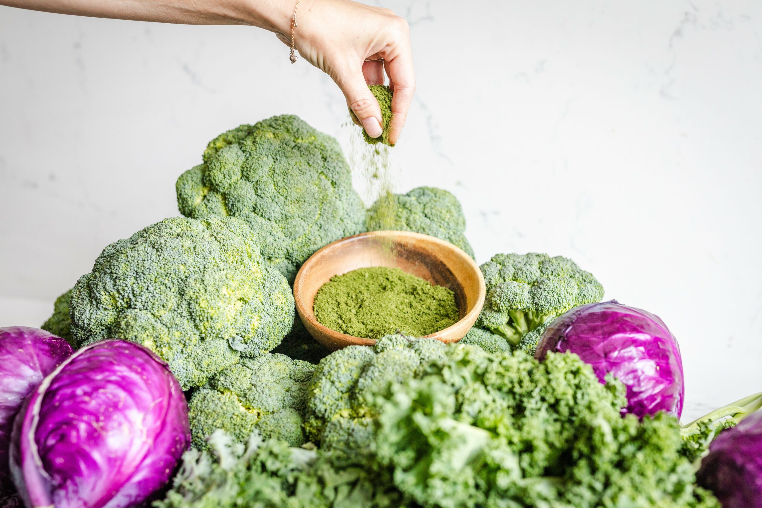 Hand sprinkling green powder over broccoli and purple cabbage on a white surface.