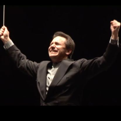 Conductor with arms raised, smiling, wearing a dark suit and white shirt, against a black background.